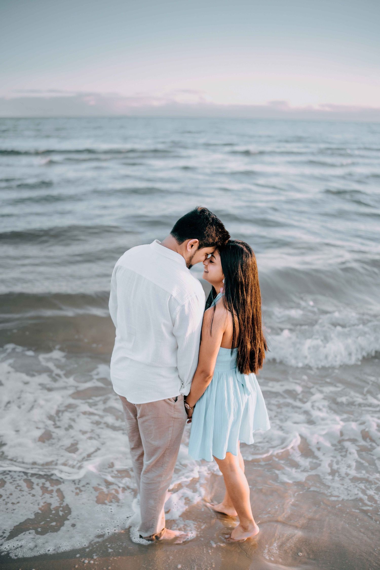 Romantic couple standing close together in ocean waves at dusk, woman in light blue dress.