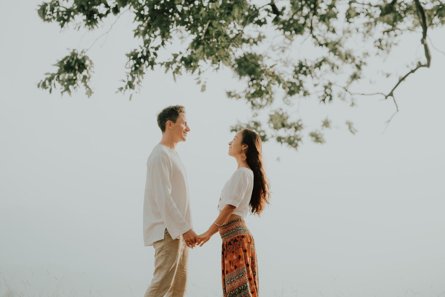 A couple holds hands and gazes at each other beneath a tree branch against a soft bright sky in a romantic outdoor setting.