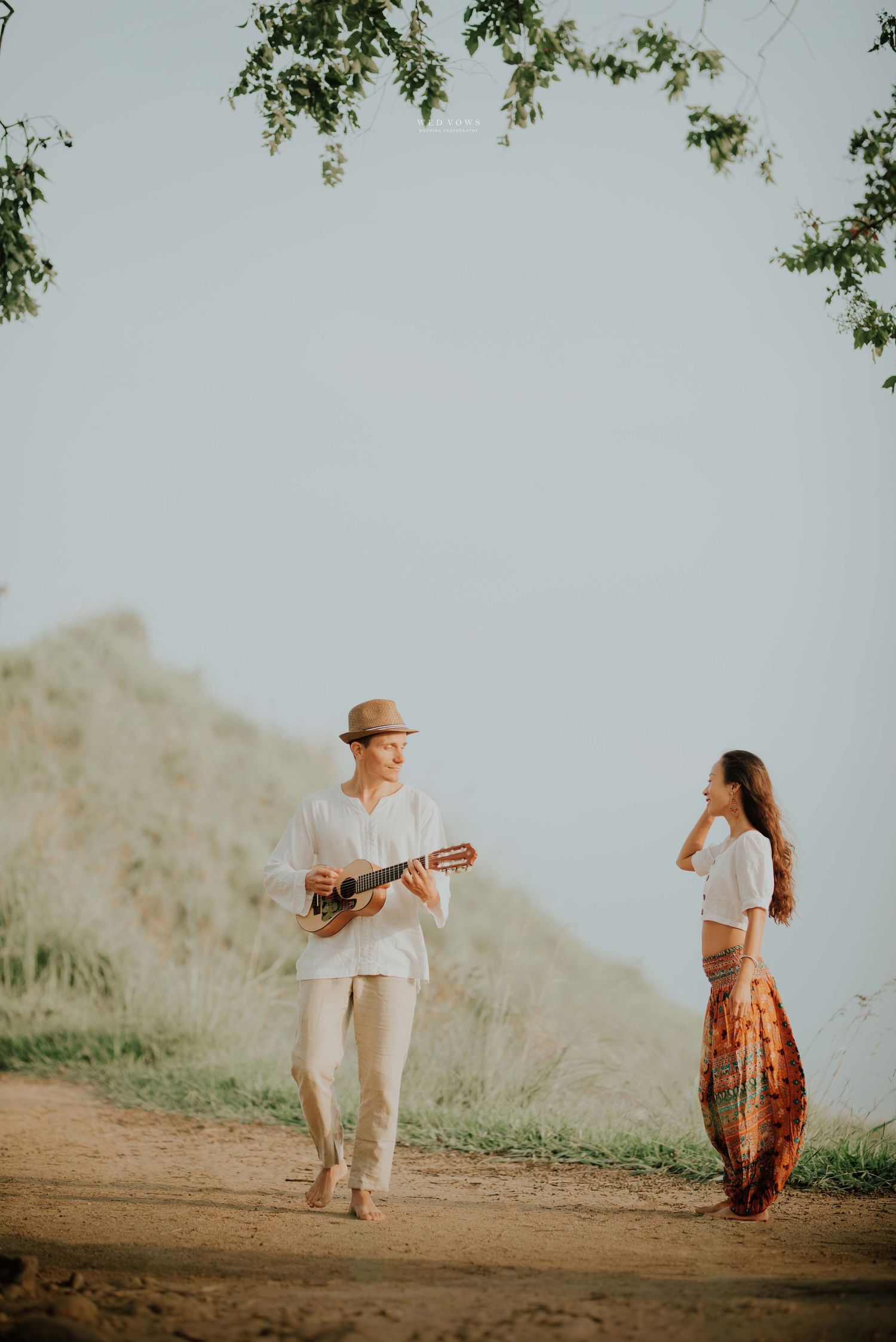 A romantic couple strolling outdoors, man playing ukulele in white outfit and hat, woman in bohemian skirt.