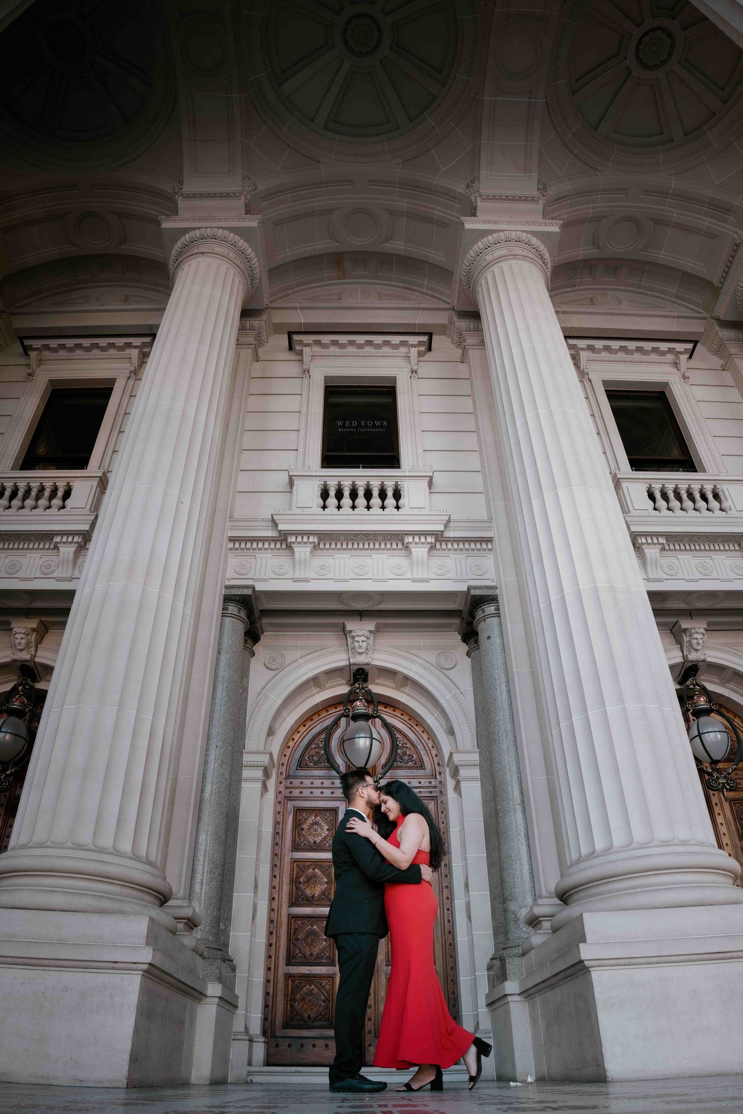 Couple embracing in red dress and black suit before grand neoclassical building with tall columns.