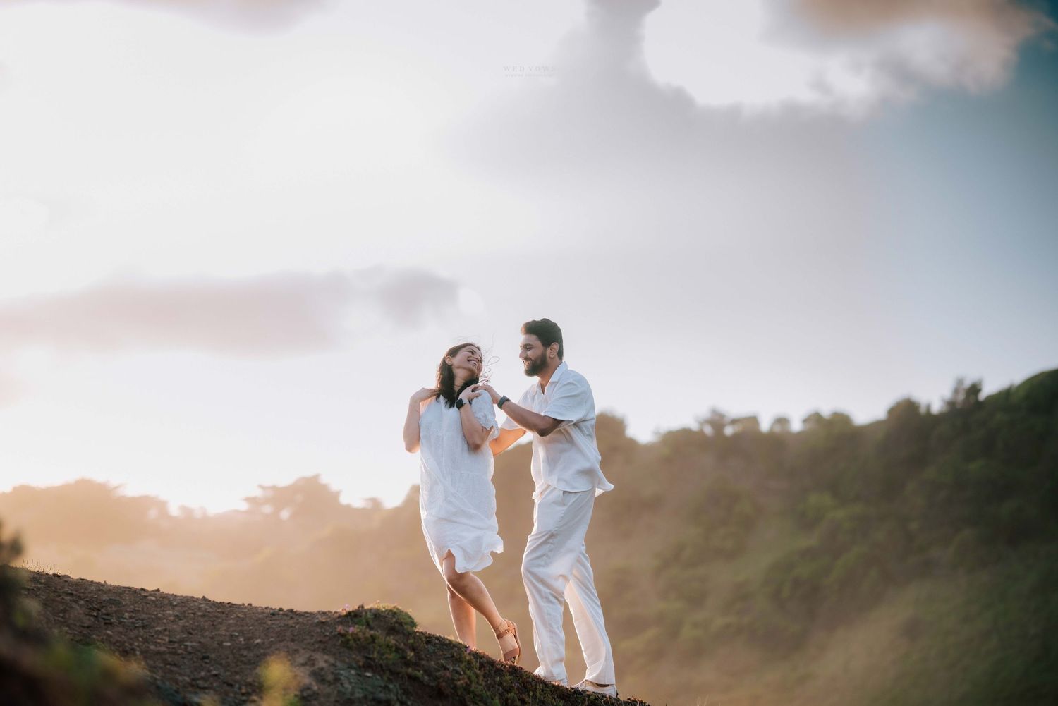 Romantic couple in matching white outfits dancing on hilltop at golden hour with lush greenery.