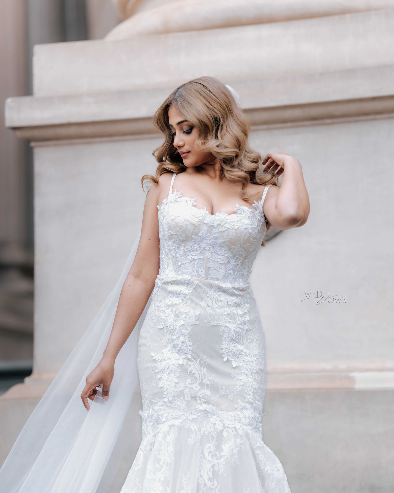 Elegant bride in fitted lace mermaid gown with veil poses beside stone column.