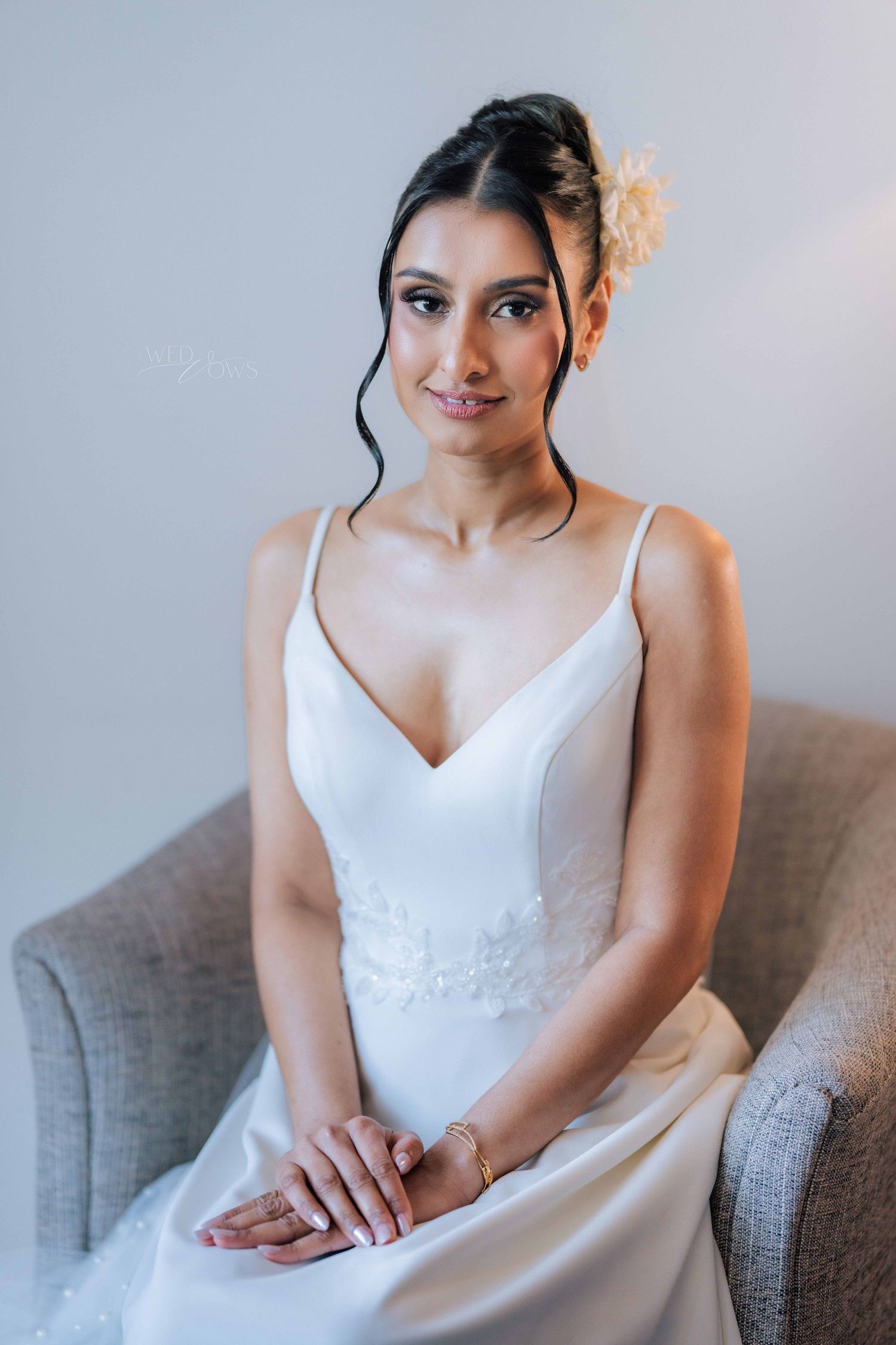 Elegant bride in white spaghetti-strap wedding dress seated in grey chair with floral hair accessory.