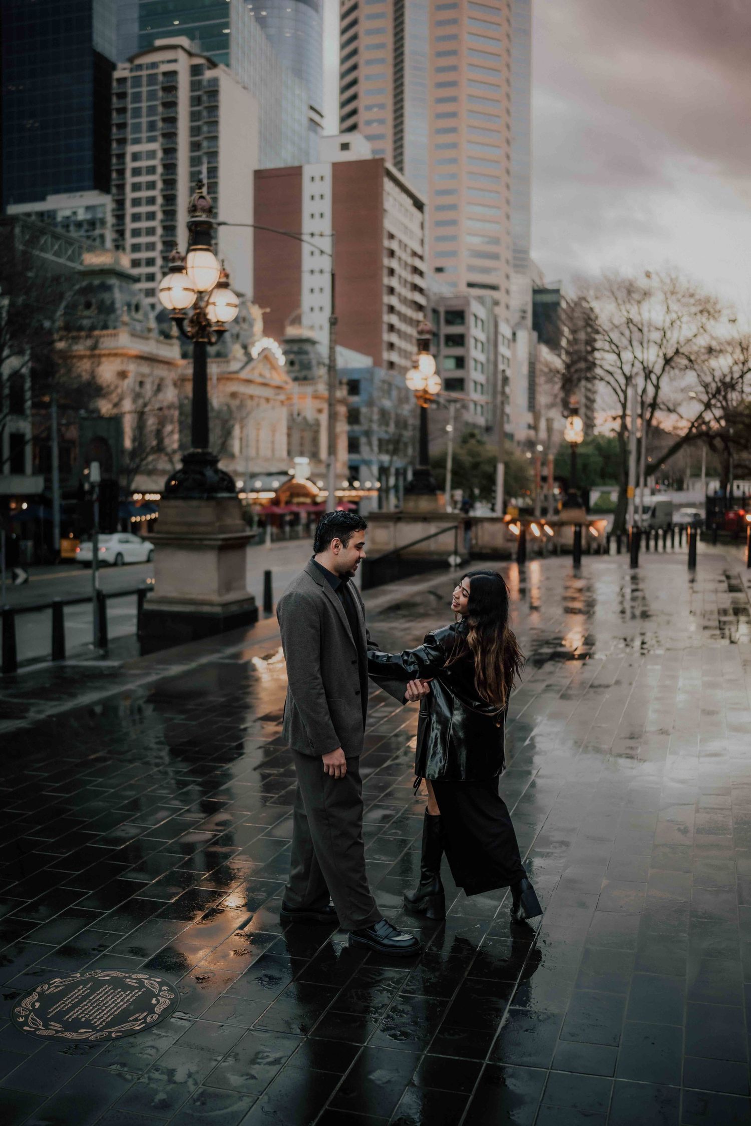 Couple holding hands facing each other on a wet city plaza with glowing street lamps.