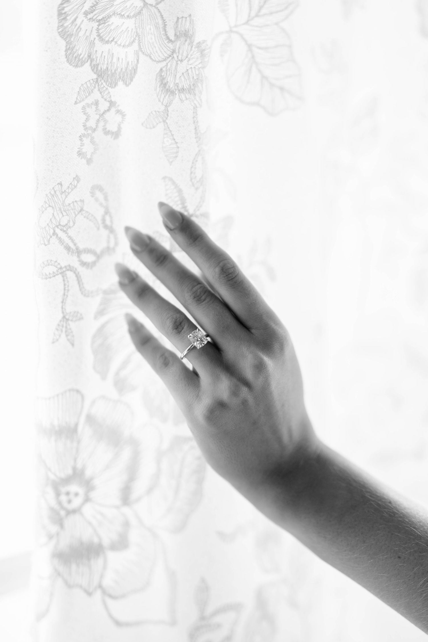 Close-up black and white photo of bride's hand with diamond engagement ring against lace dress.