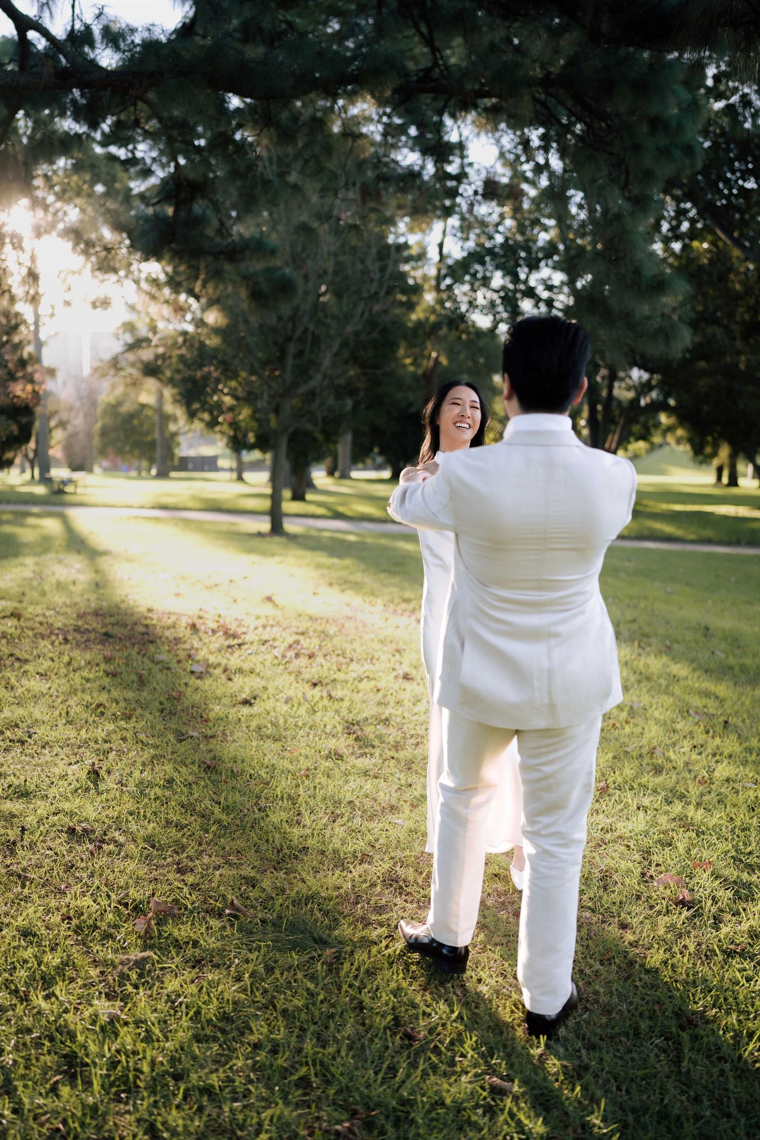 Couple in white outfits sharing a romantic moment in a sunlit park with lush green trees.
