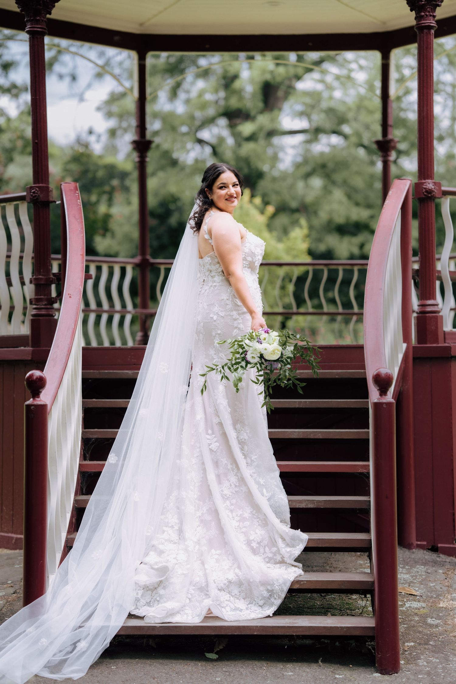 Bride in lace gown with veil poses on gazebo stairs holding white bouquet.