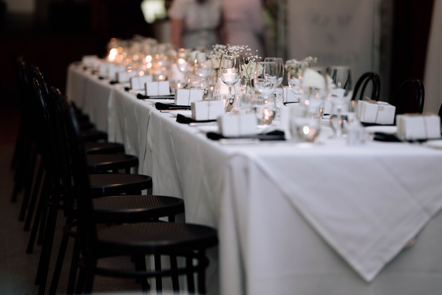 Long elegantly set wedding reception table with white linens, dark chairs, candles, and glassware.