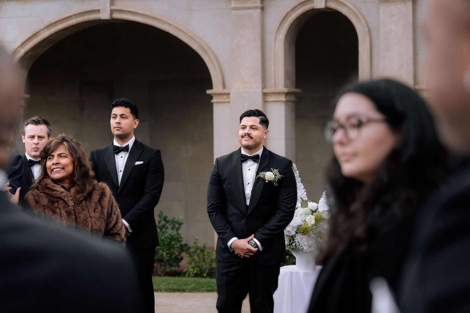 Groom watching bride approach during outdoor wedding ceremony at historic venue.