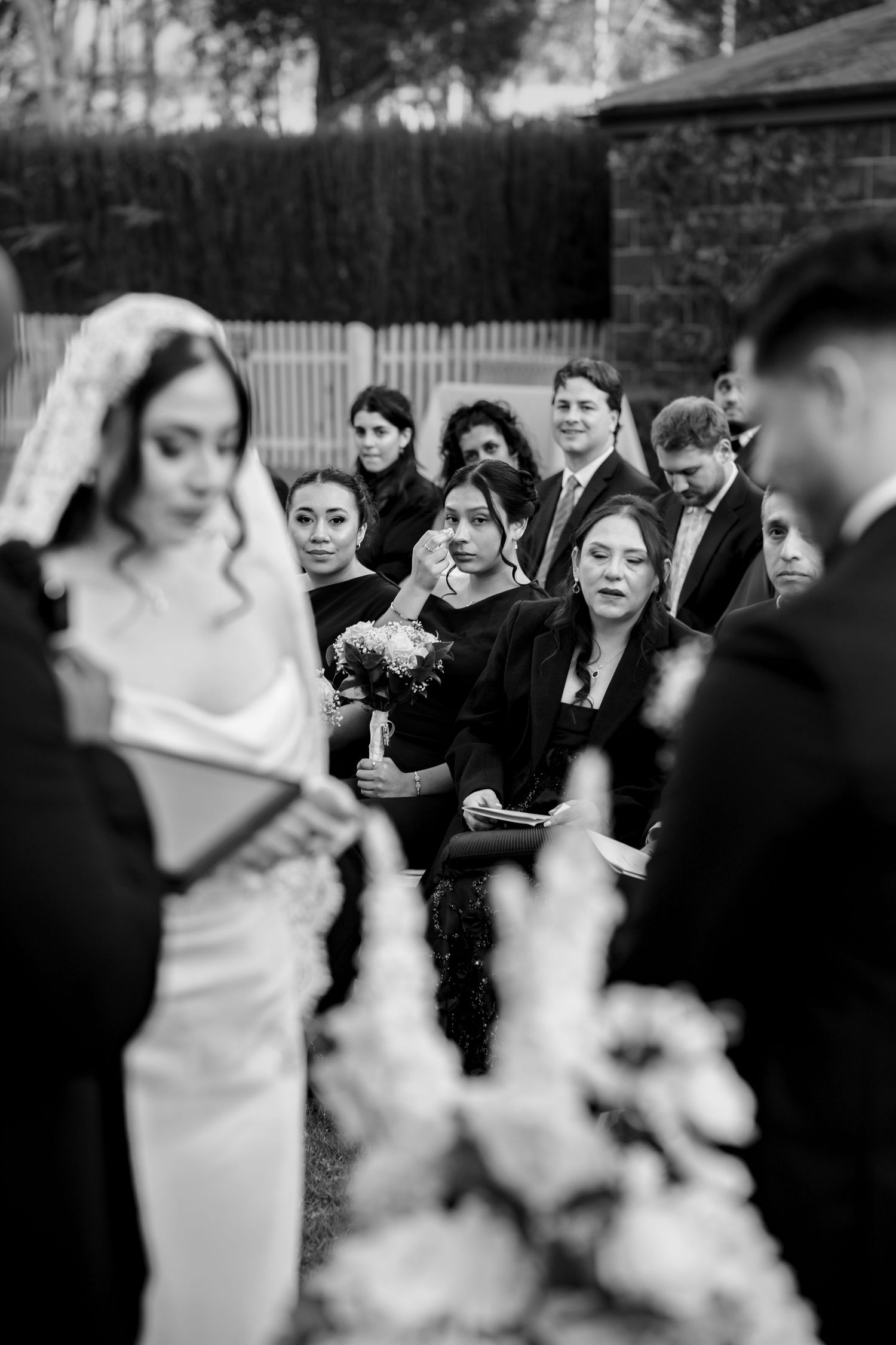Black and white photo of emotional wedding guests watching outdoor ceremony as bride stands at altar.