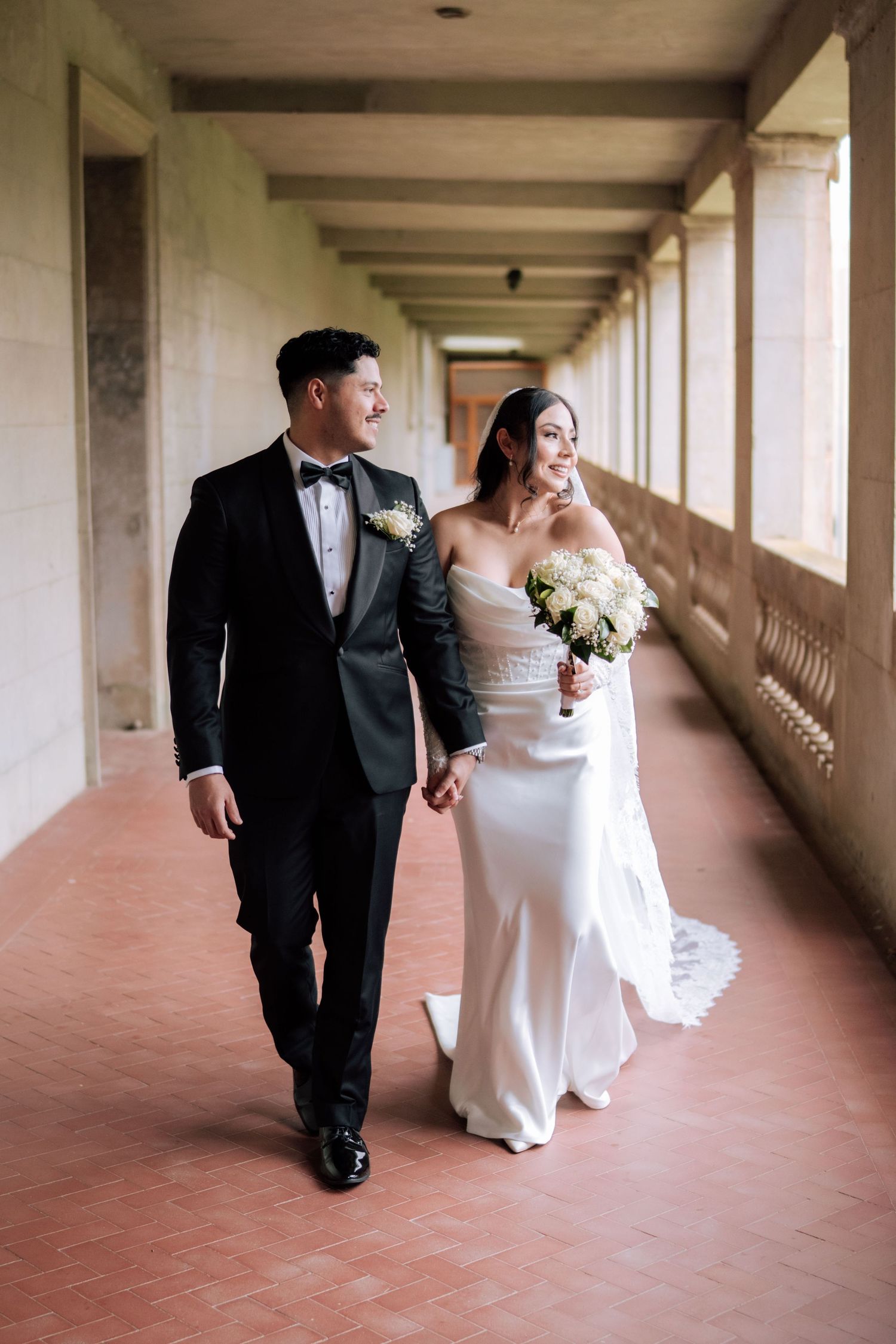 A bride and groom walk hand in hand through an elegant columned hallway, she in a white gown holding flowers, he in a black tuxedo.