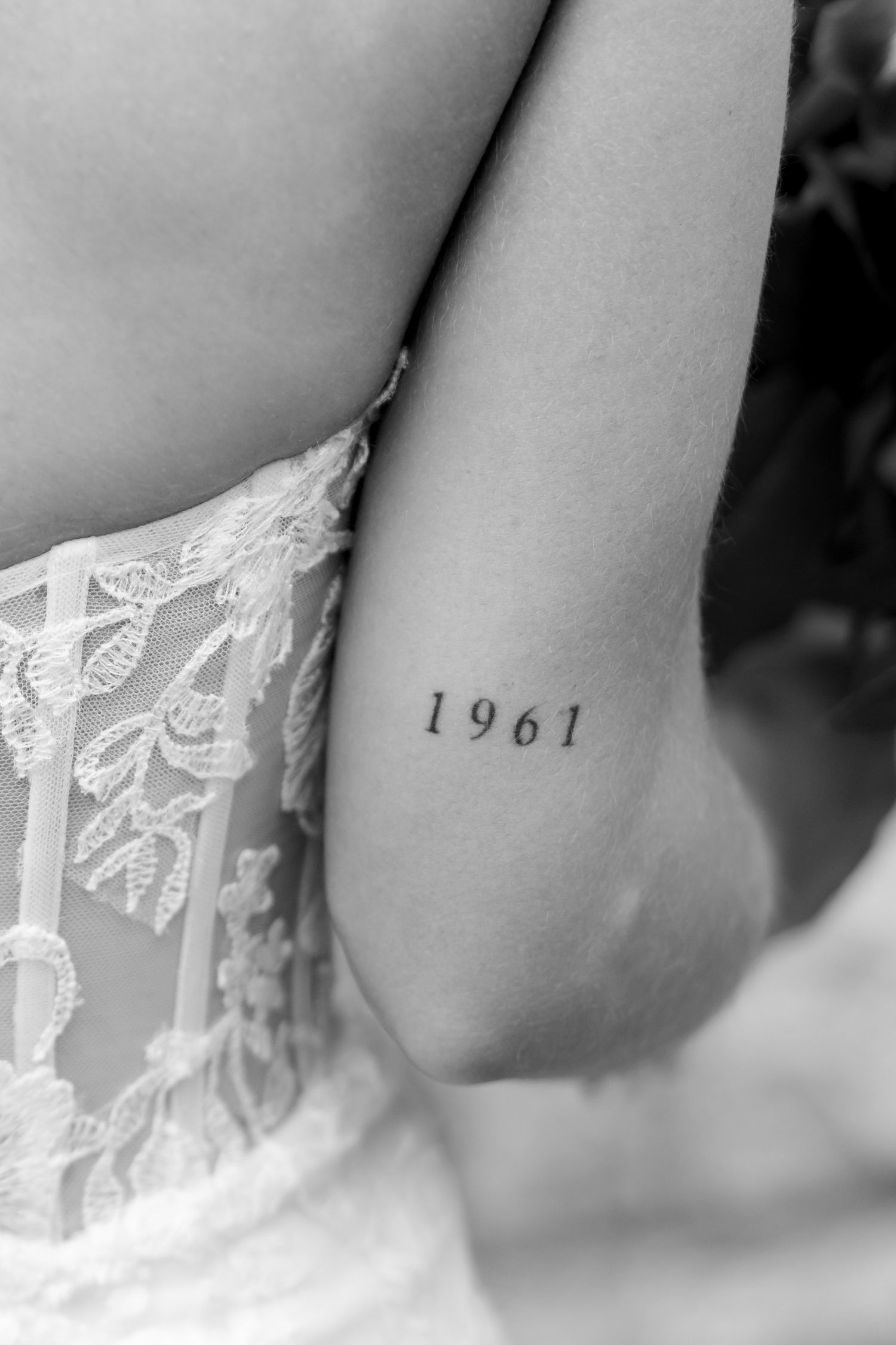 Close-up black and white photo of bride with small 1961 tattoo on her arm beside lace wedding dress.