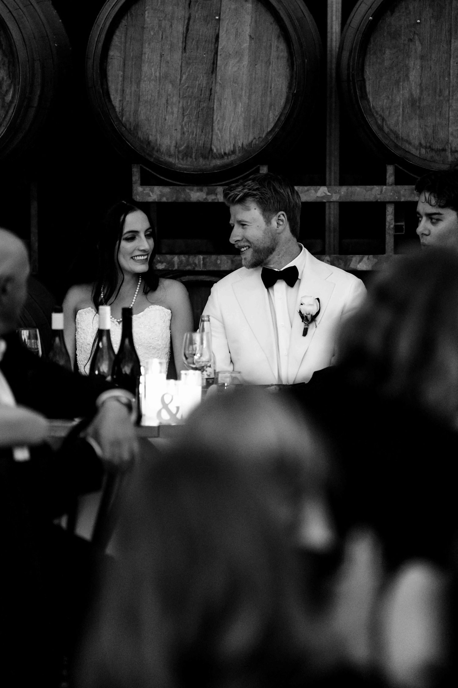 Black and white photo of bride and groom smiling at wedding reception with wine barrels in background.