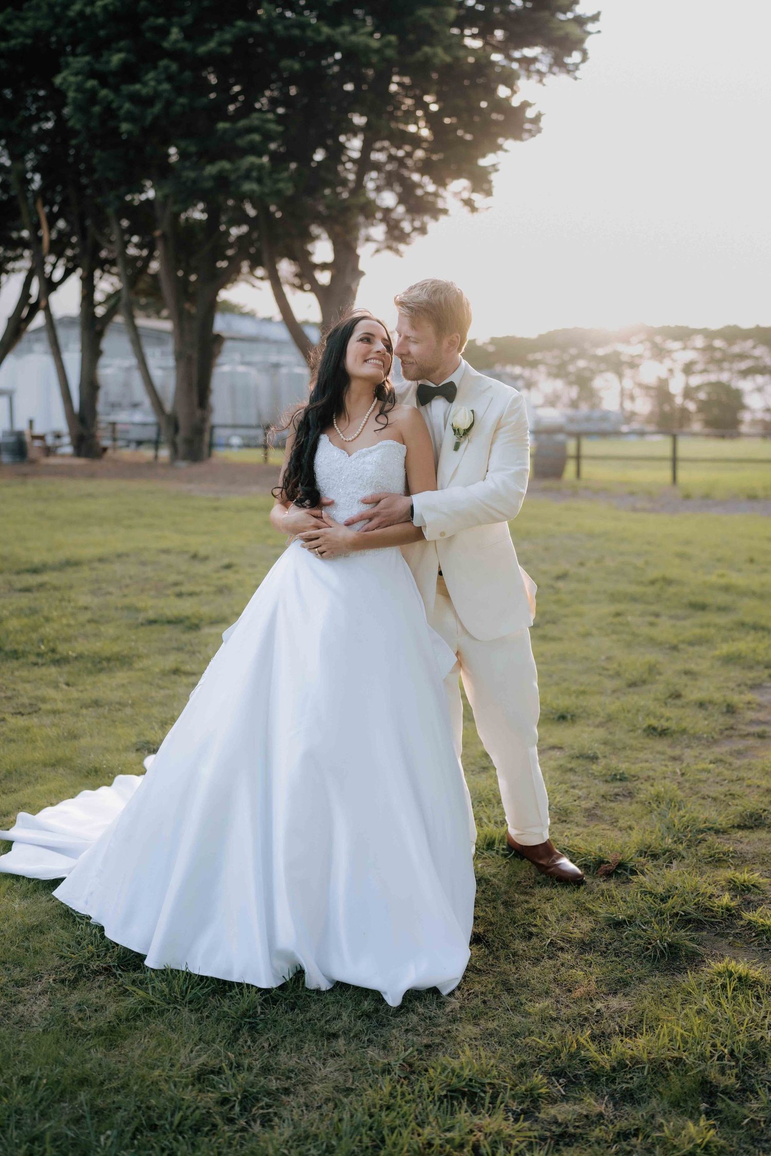 Groom in cream suit embraces bride in strapless white ballgown at golden hour in a lush green park setting.