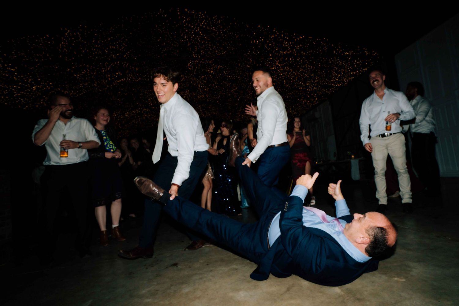 Groomsmen dance energetically at a wedding reception, with one guest lying on the dance floor during celebrations.