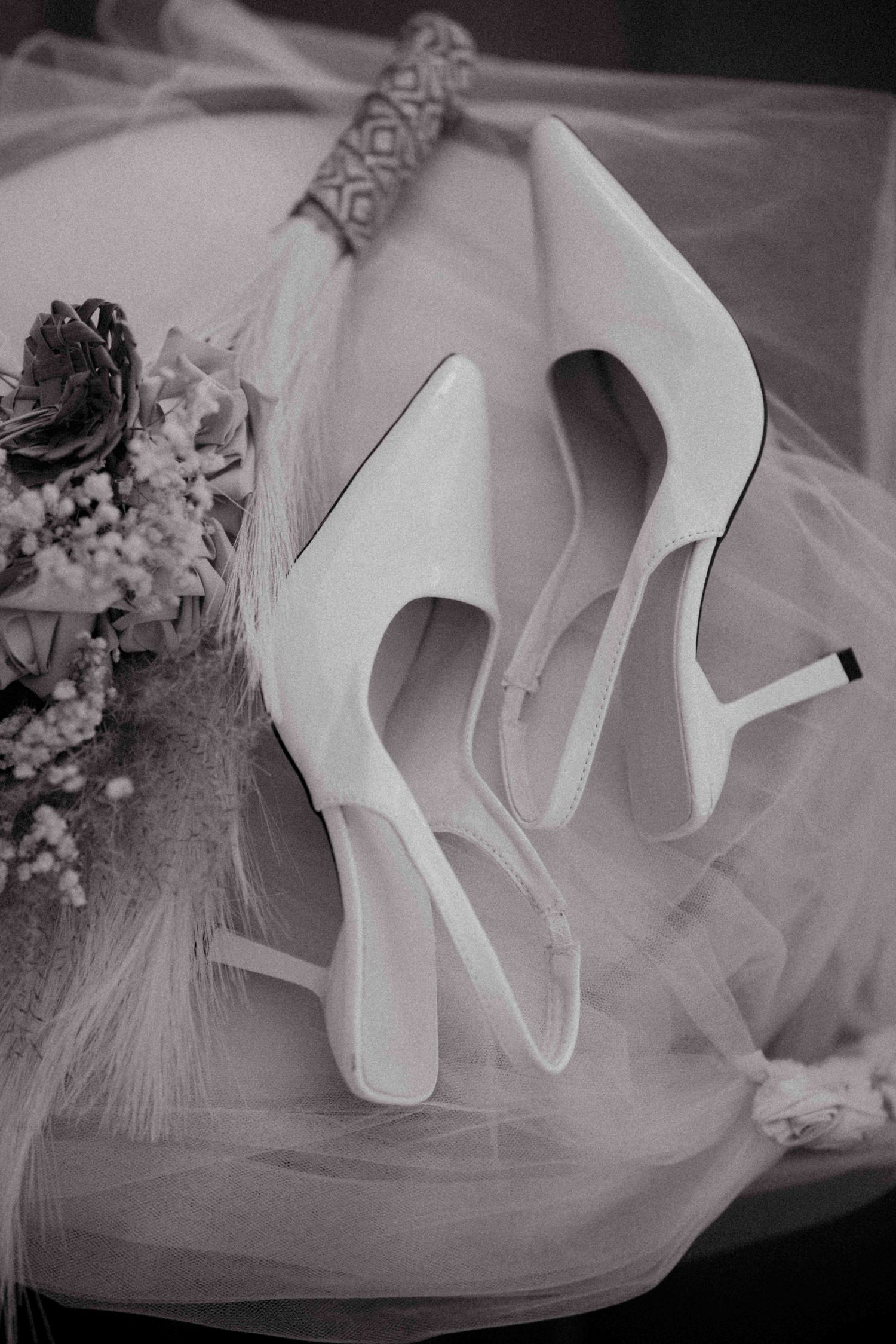 Black and white photo of white bridal heels on tulle veil beside a dried flower bouquet.