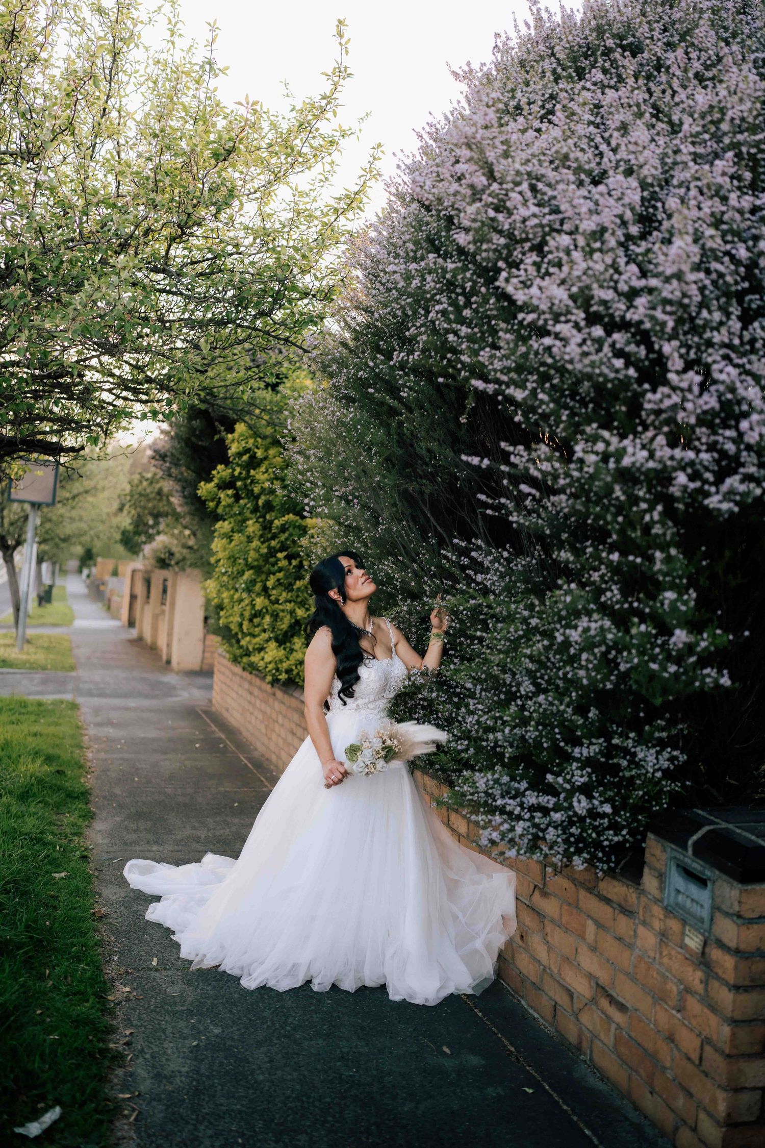 Beautiful bride in white tulle wedding gown standing beside blooming pink flowering bushes outdoors.