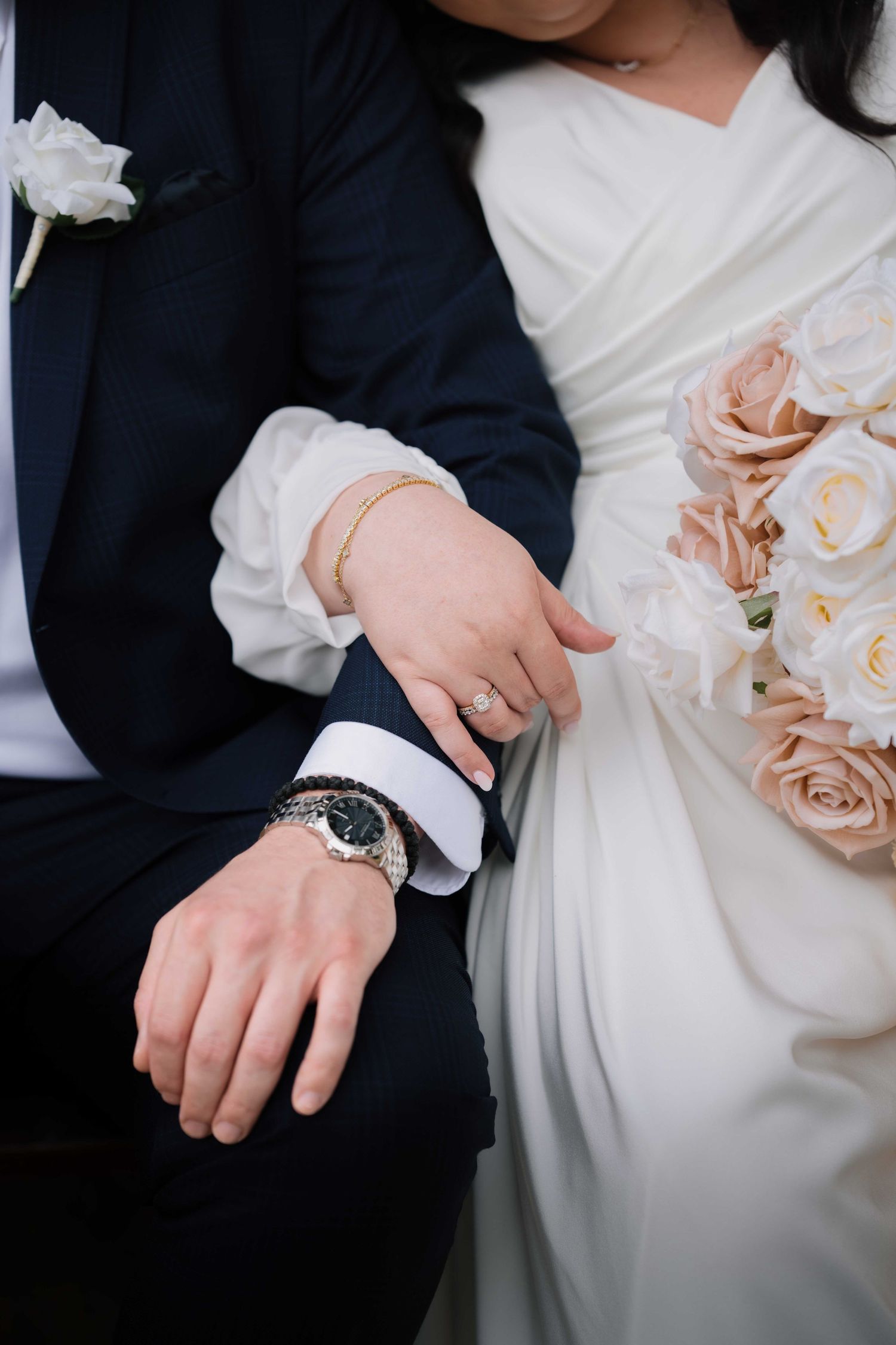 Close-up of bride and groom hands showing diamond ring, gold bracelet, and watch with blush rose bouquet.