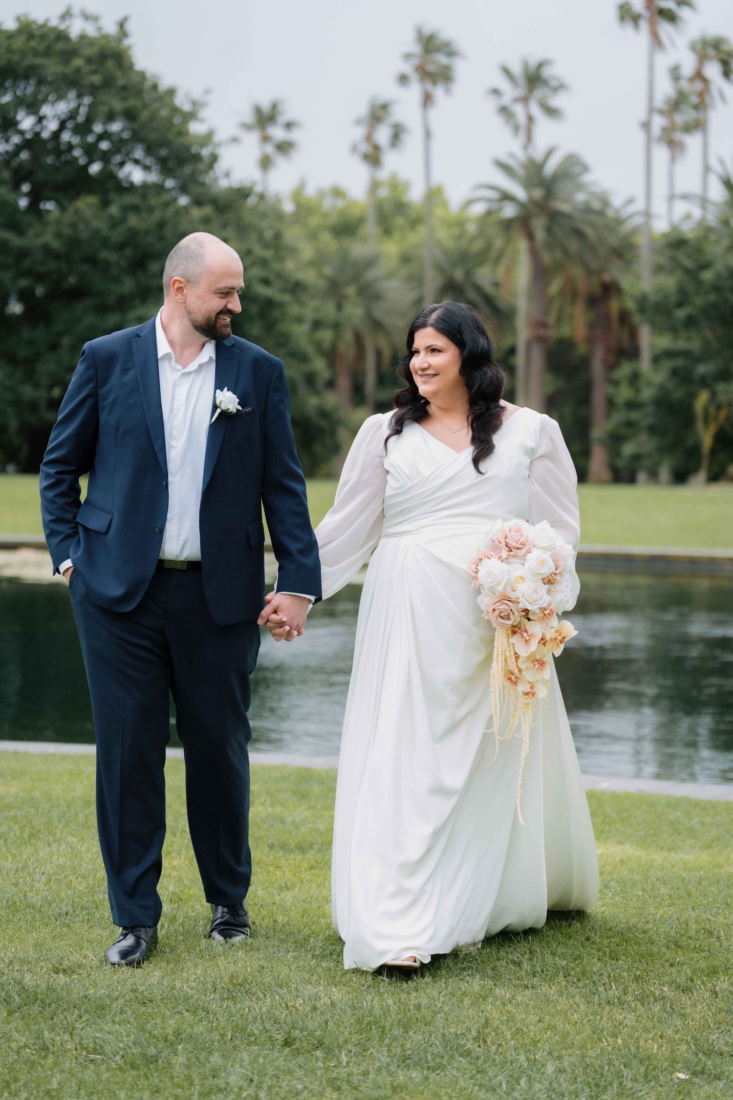 Bride and groom stroll hand in hand on green lawn with palm trees and pond backdrop.
