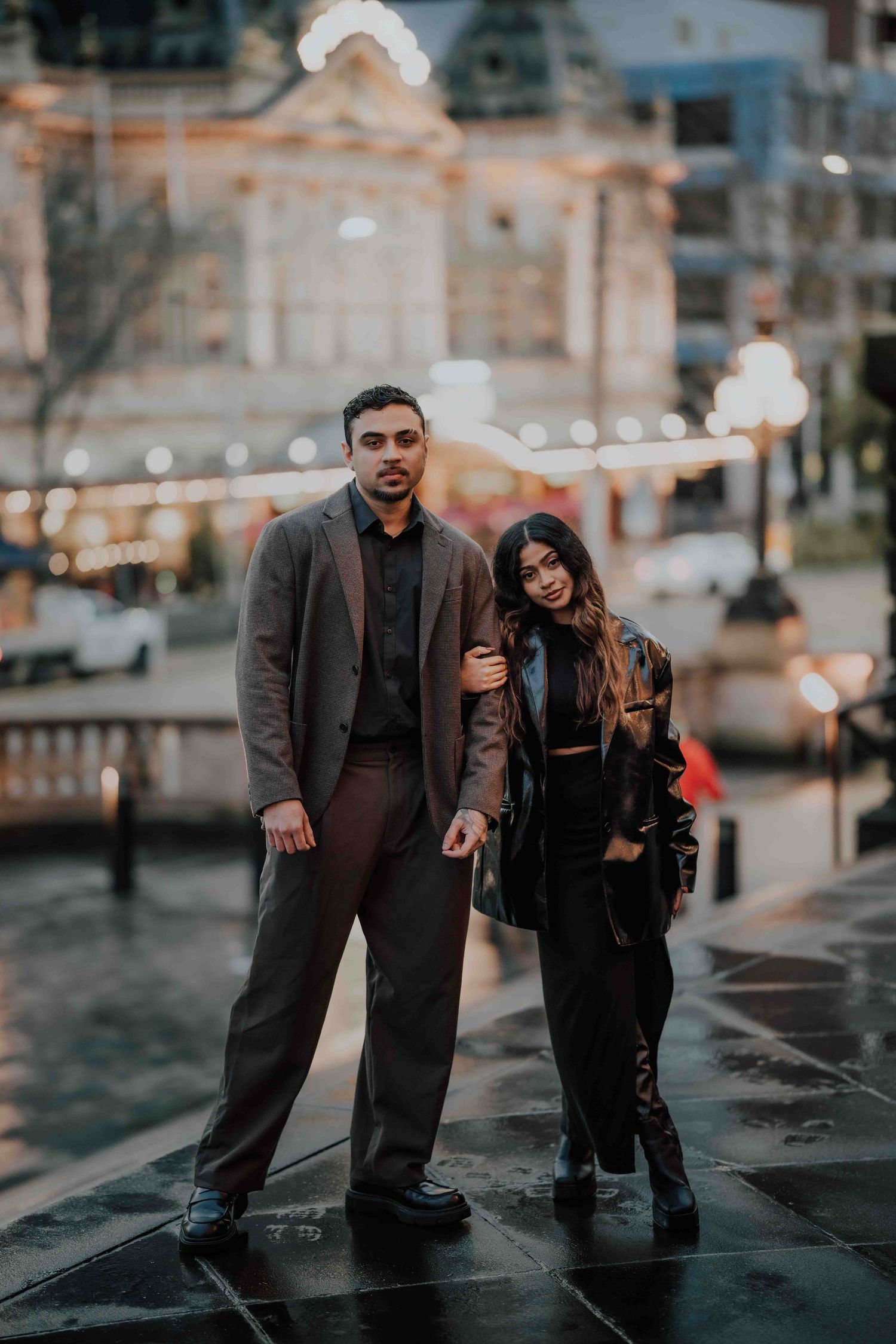Stylish couple posing together on wet pavement with illuminated city buildings at dusk.