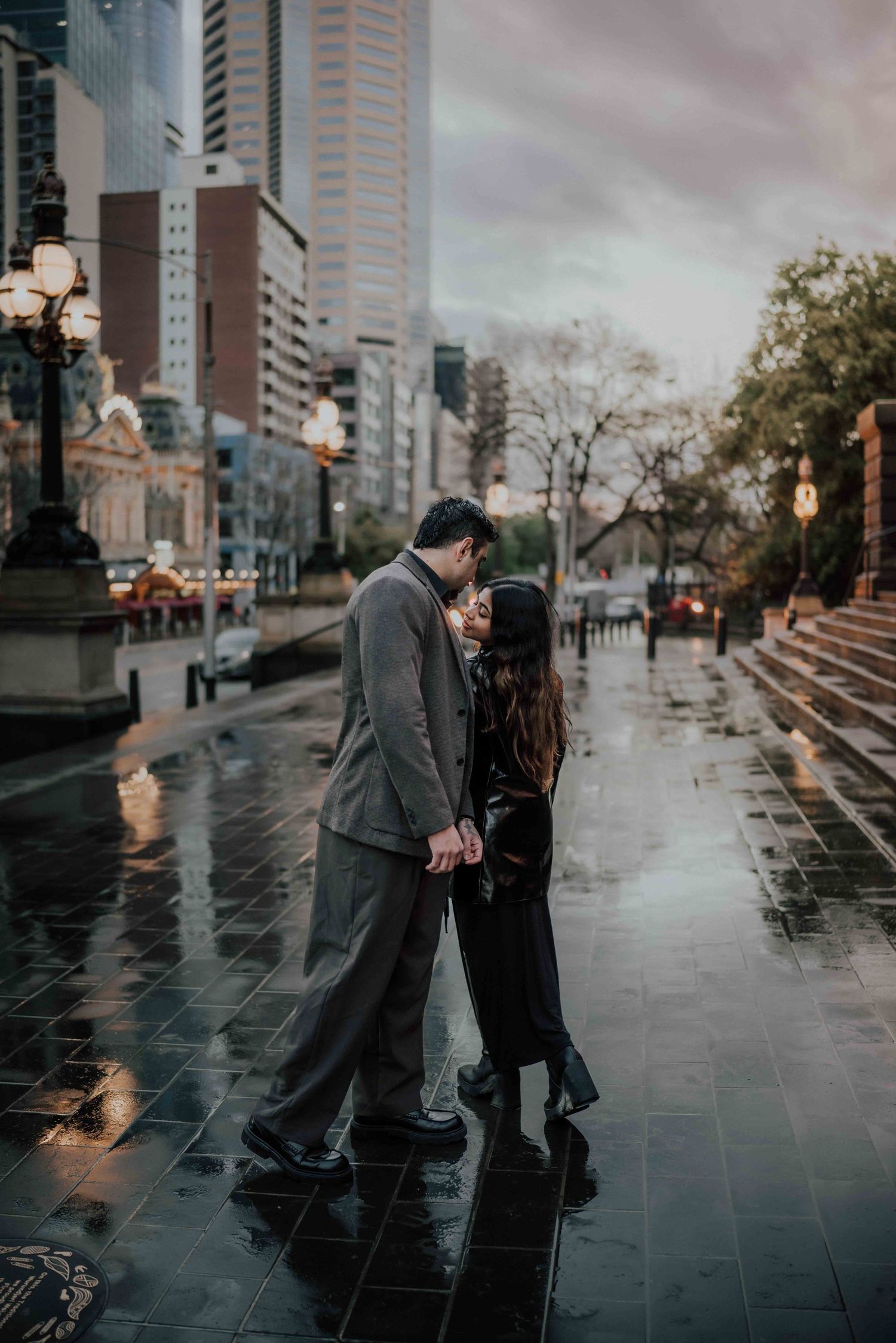 Couple kissing on a rainy city street at dusk with urban skyline in the background.