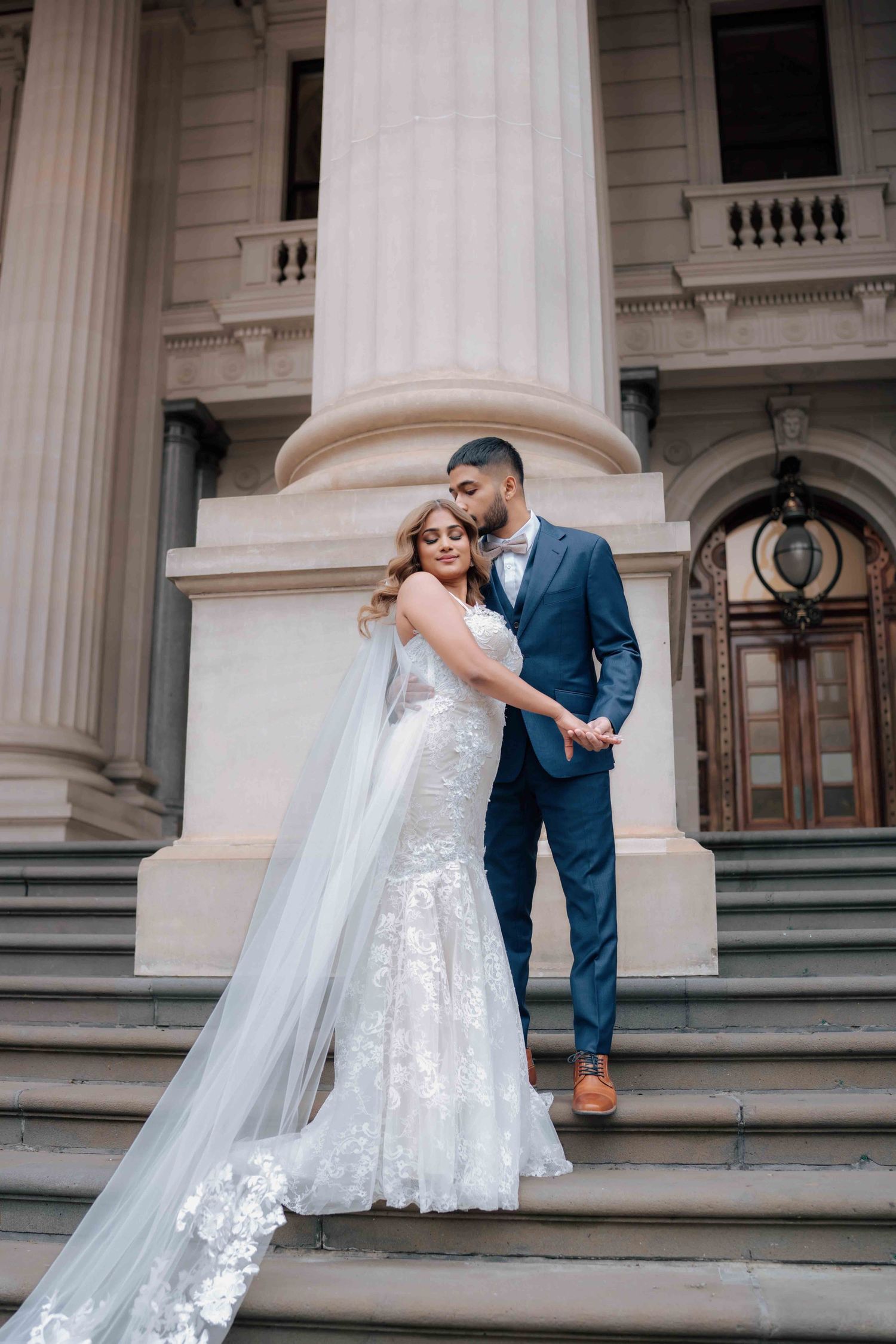 Groom in navy suit embracing bride in lace mermaid gown on steps of classical building.