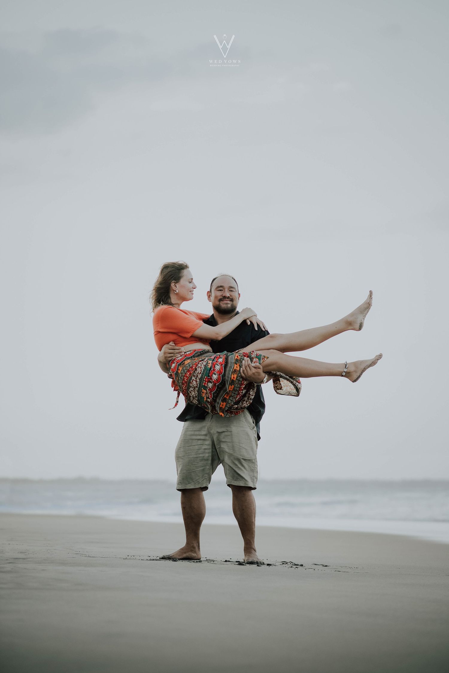 Man carrying woman in his arms on a sandy beach, both laughing joyfully under a cloudy sky.