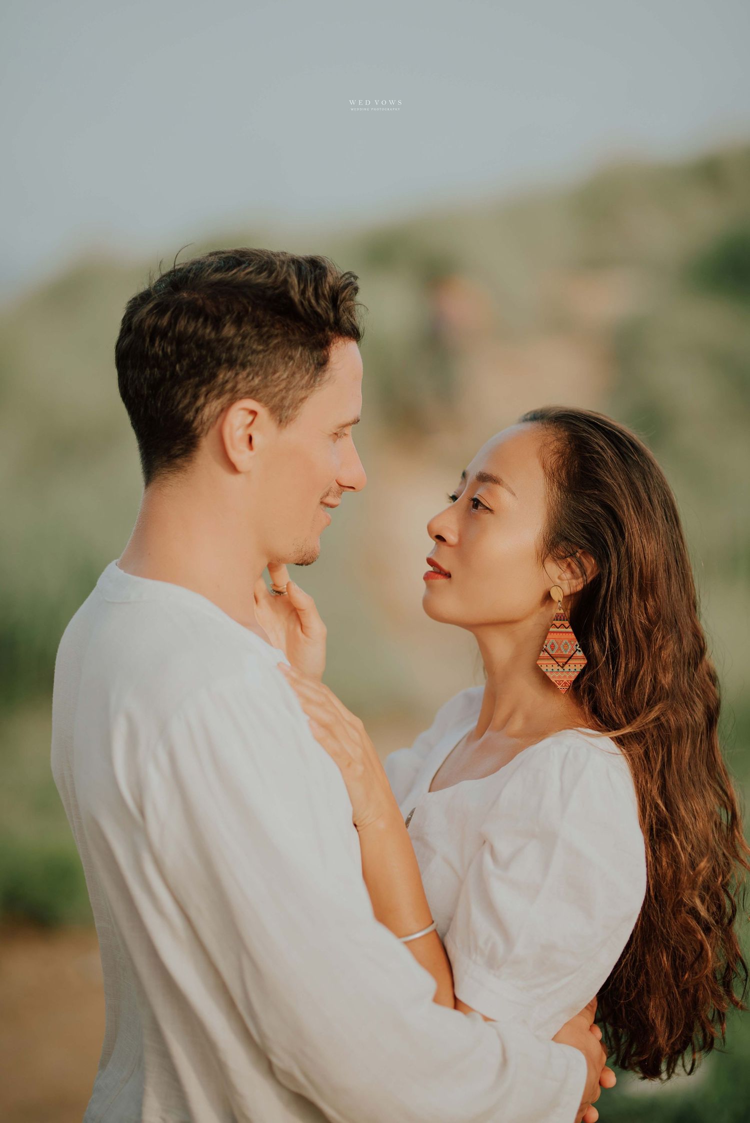 Romantic couple in white outfits gazing into each other's eyes at golden hour.