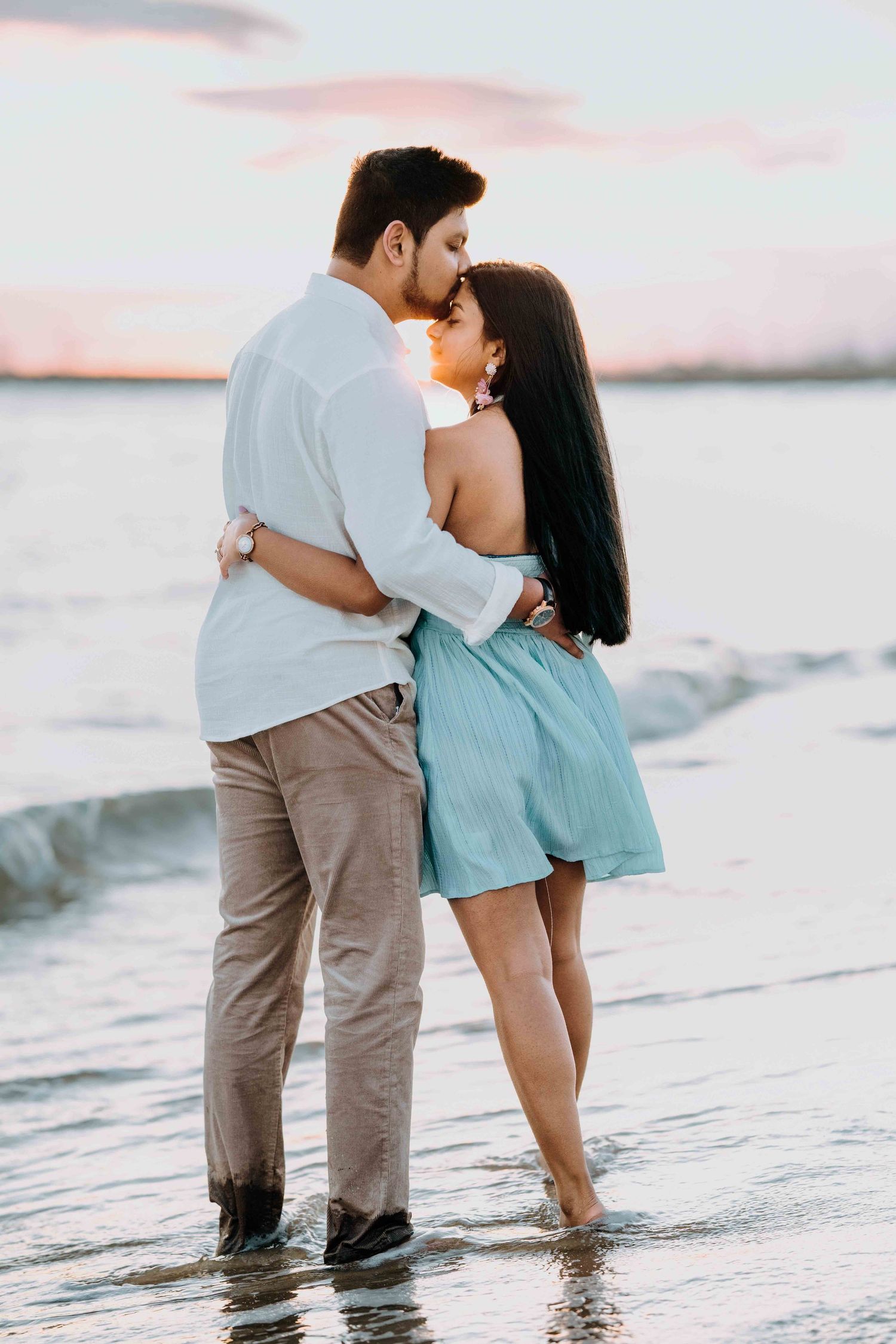Romantic couple kissing at sunset on a beach, woman in light blue dress embracing man in white shirt.