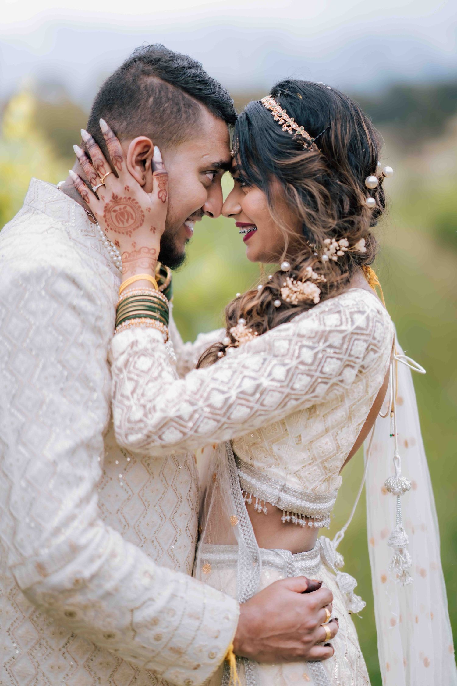 Indian bride and groom in white embroidered outfits touching foreheads, bride wearing mehndi and gold jewelry outdoors.