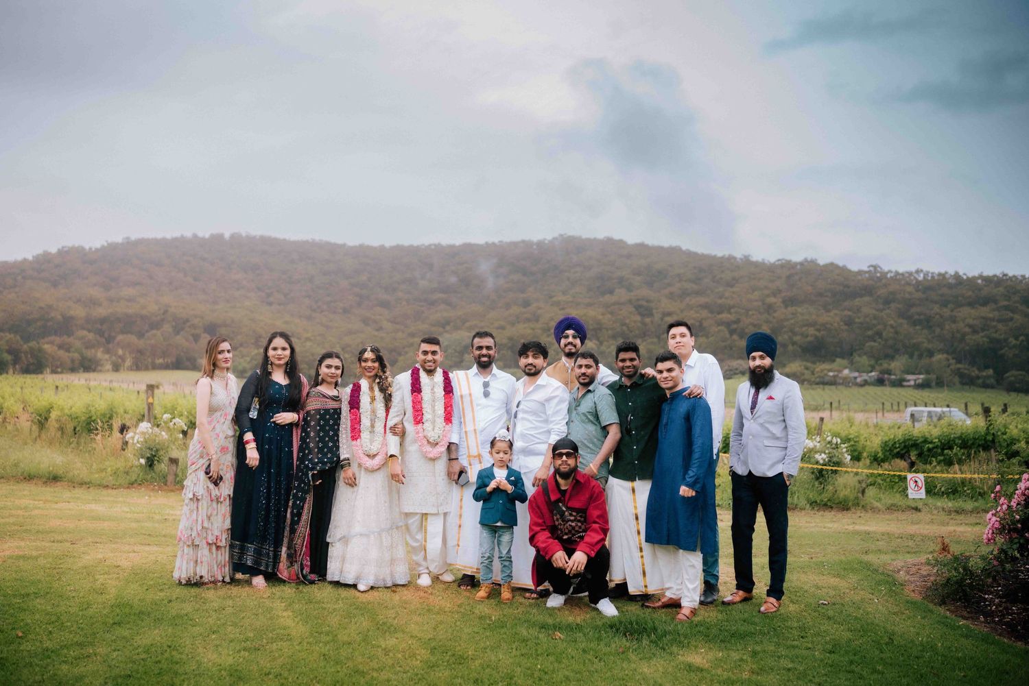 Indian wedding group portrait with bride and groom surrounded by family and friends on a green vineyard lawn.