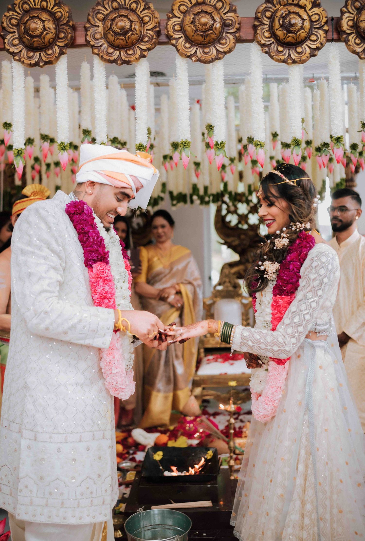 Smiling Hindu bride and groom exchanging garlands at decorated wedding mandap with floral backdrop.