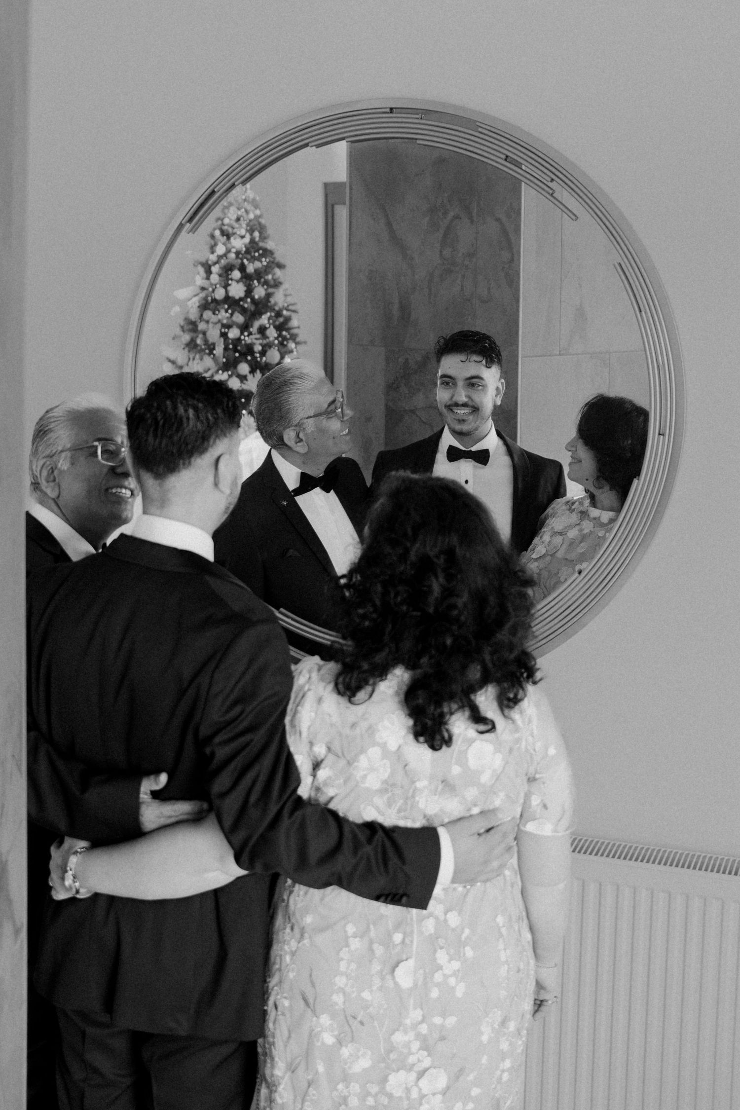 Groom sharing joyful moment with family reflected in round mirror during wedding preparations.