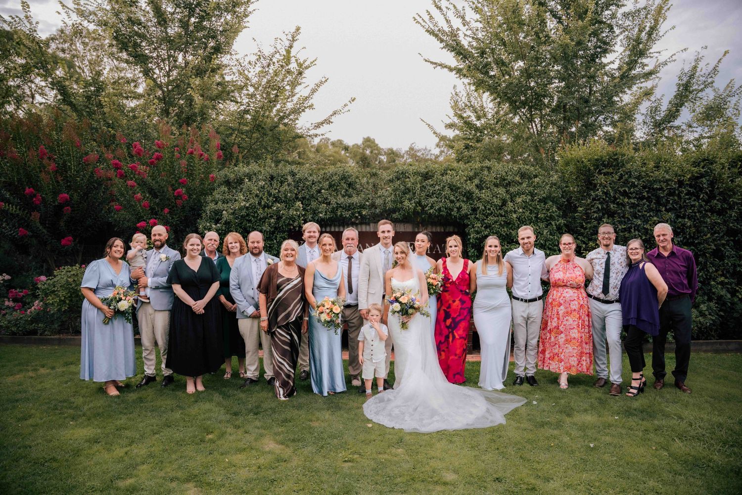 Extended family wedding portrait on grass with bride in white gown and groom surrounded by guests outdoors.