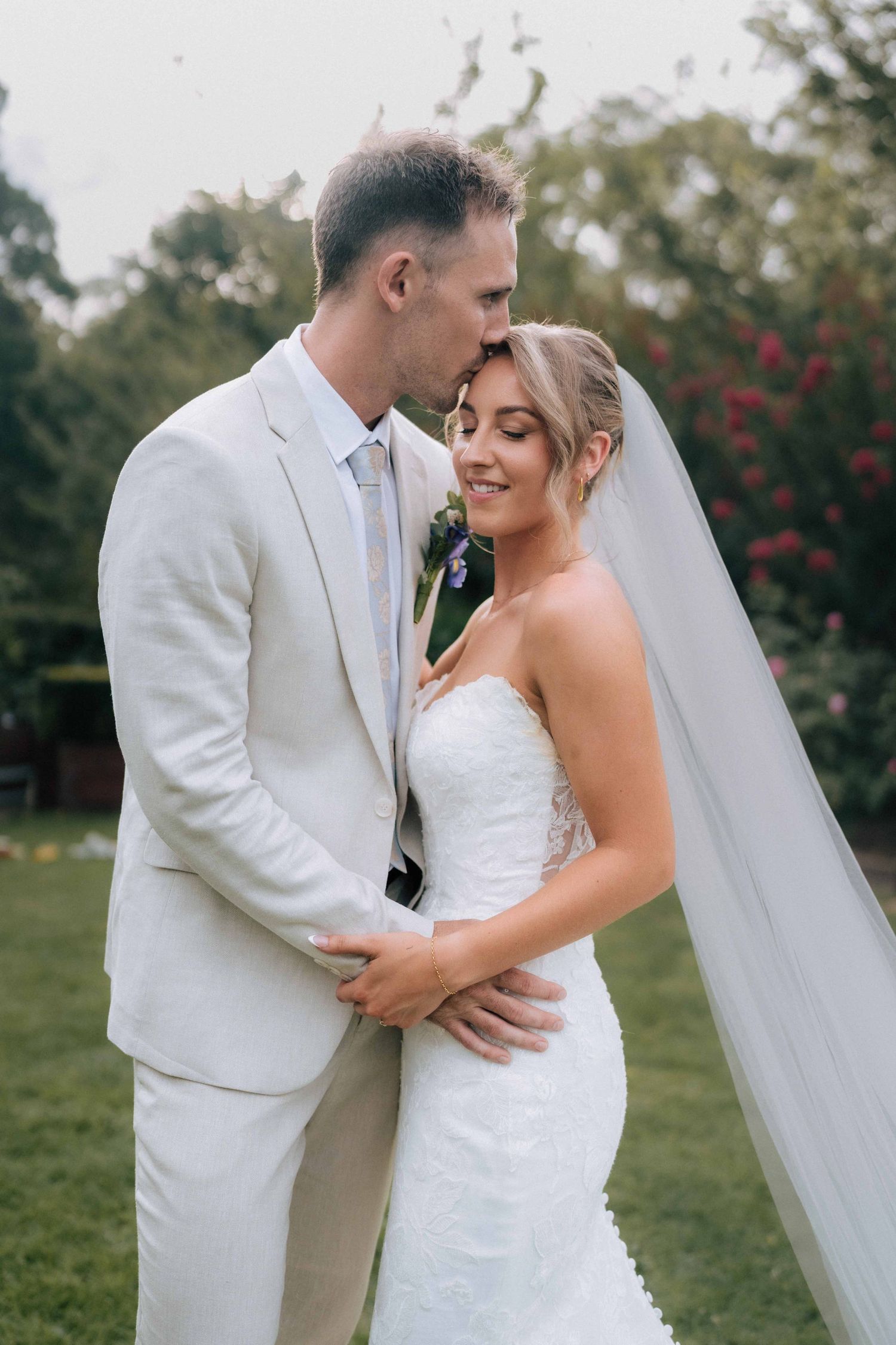 Bride and groom embracing in garden, groom in beige suit kissing brides head, bride in lace wedding gown with veil.