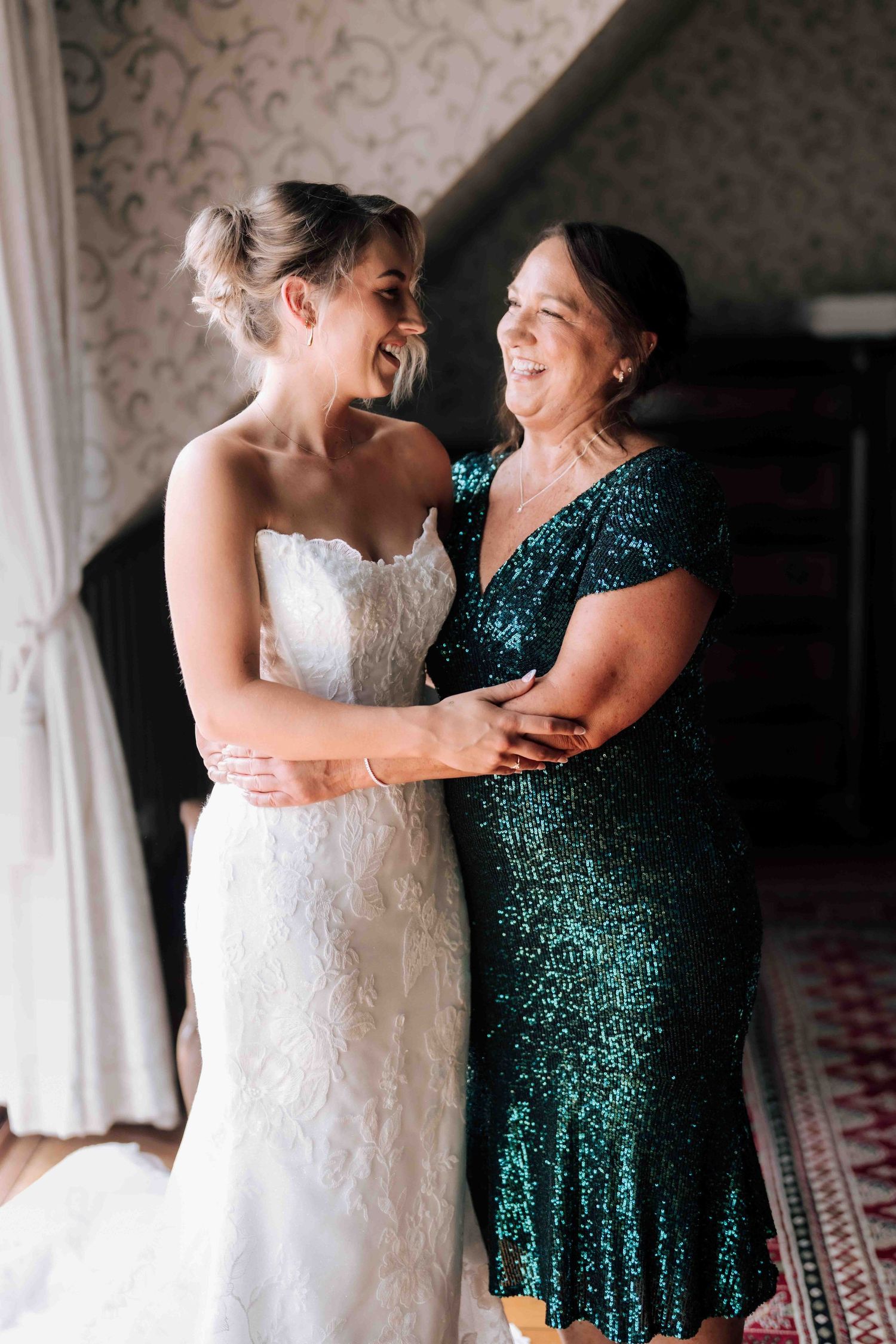 Joyful bride and mother sharing a laugh and embrace during wedding morning preparations.