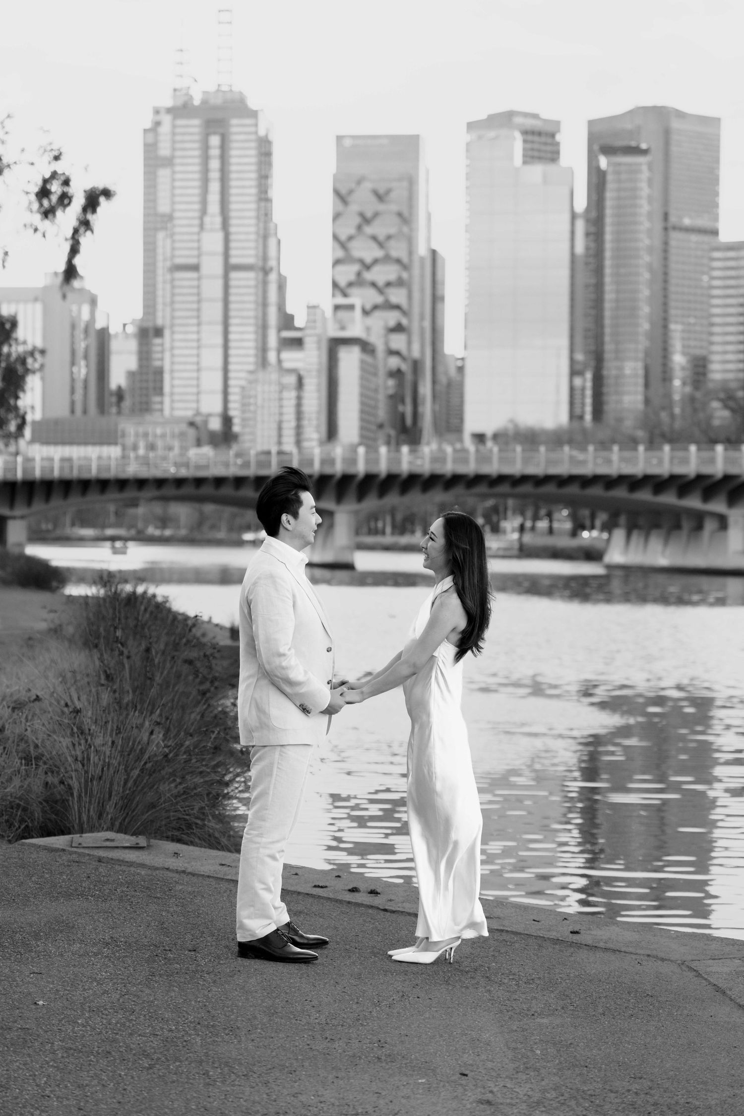 Black and white photo of a couple holding hands by a river with a city skyline and bridge in the background.