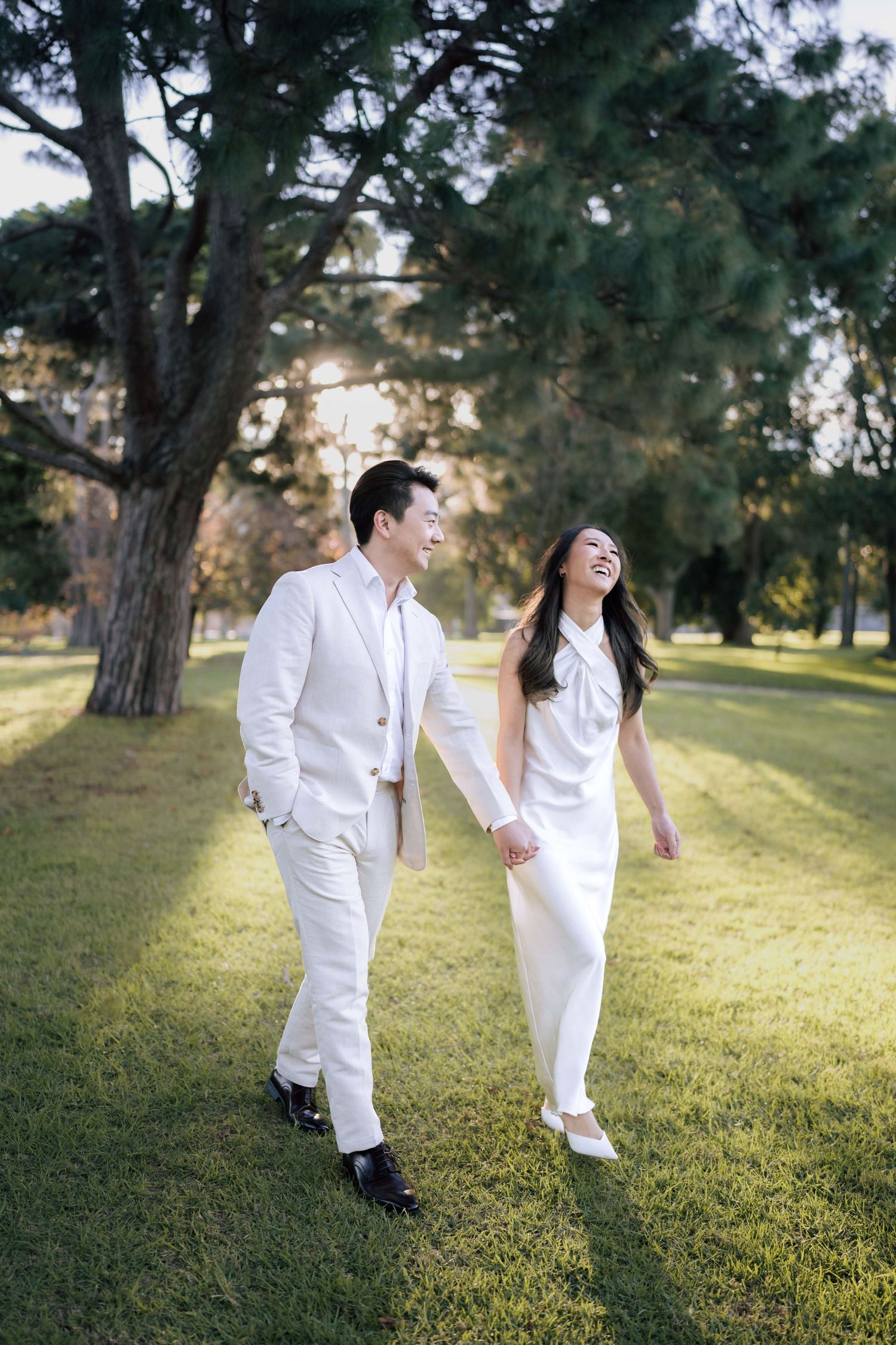 Happy couple in white outfits holding hands and laughing while walking on green grass in a sunny park.