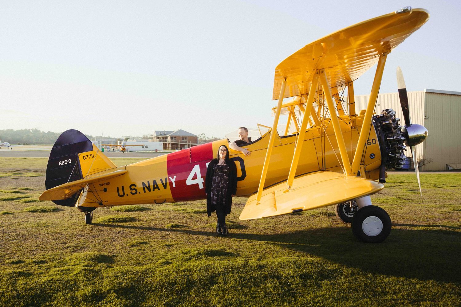 Yellow U.S. Navy biplane on grass airfield with two people standing beside it at sunset.