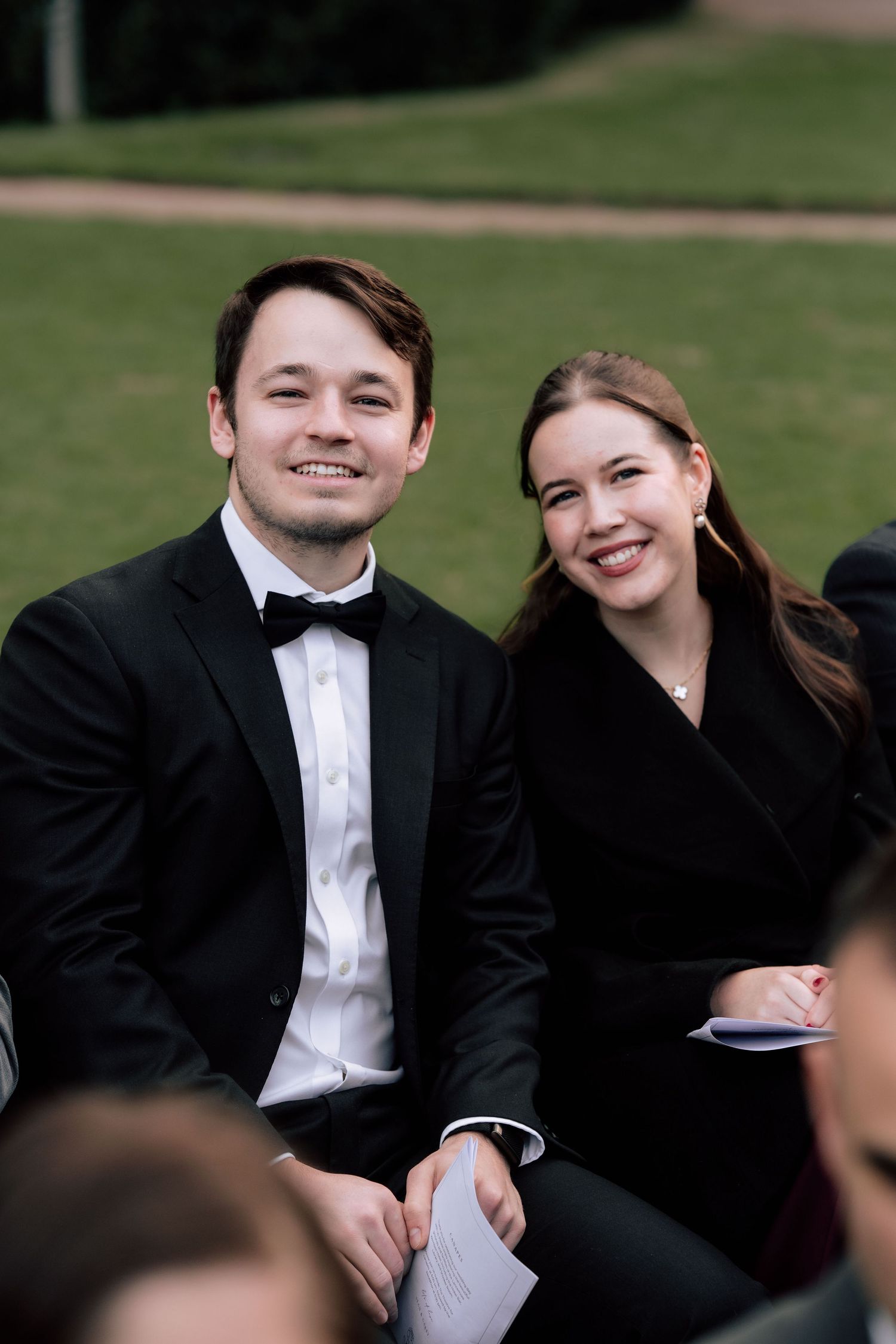 Smiling wedding guests, man in black tuxedo and woman in black dress, seated at outdoor ceremony.