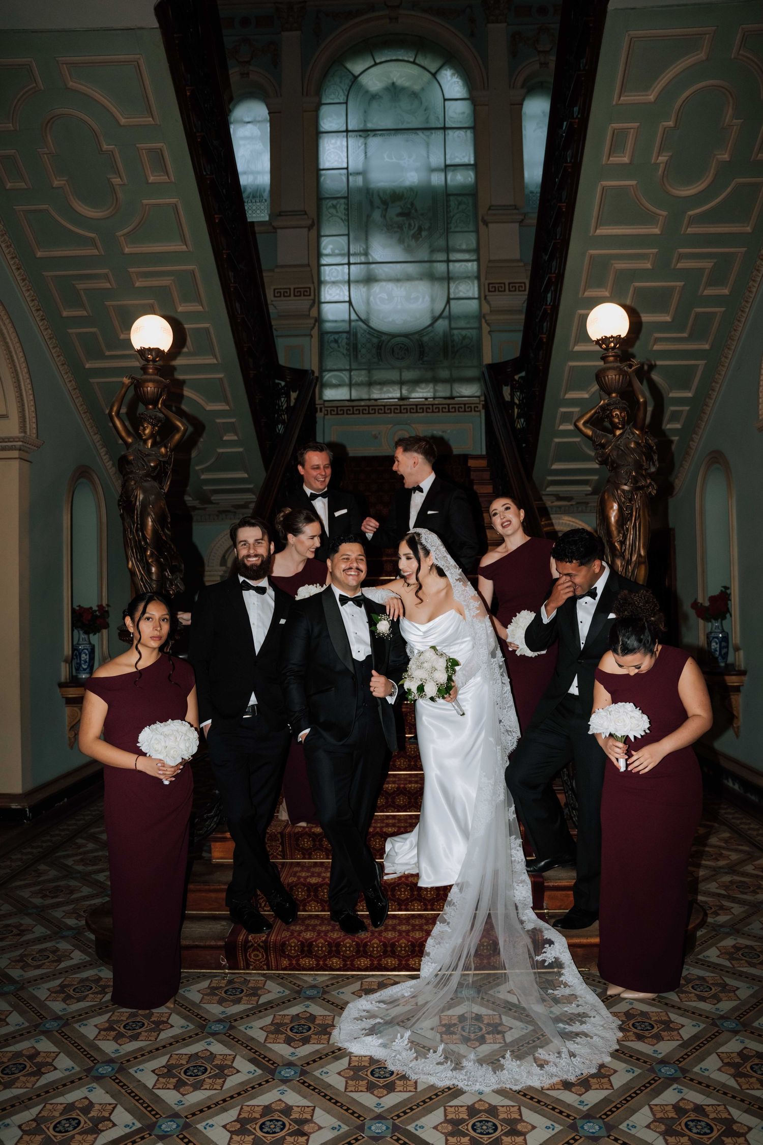 Wedding party in black tuxedos and burgundy bridesmaid dresses pose on ornate staircase in elegant venue.