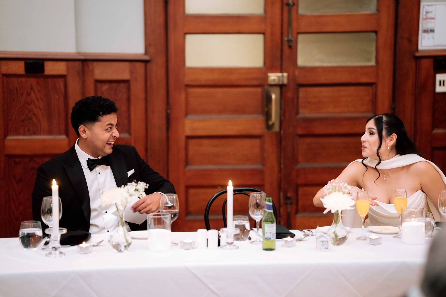 Bride and groom share a smile at sweetheart table during elegant candlelit wedding reception.