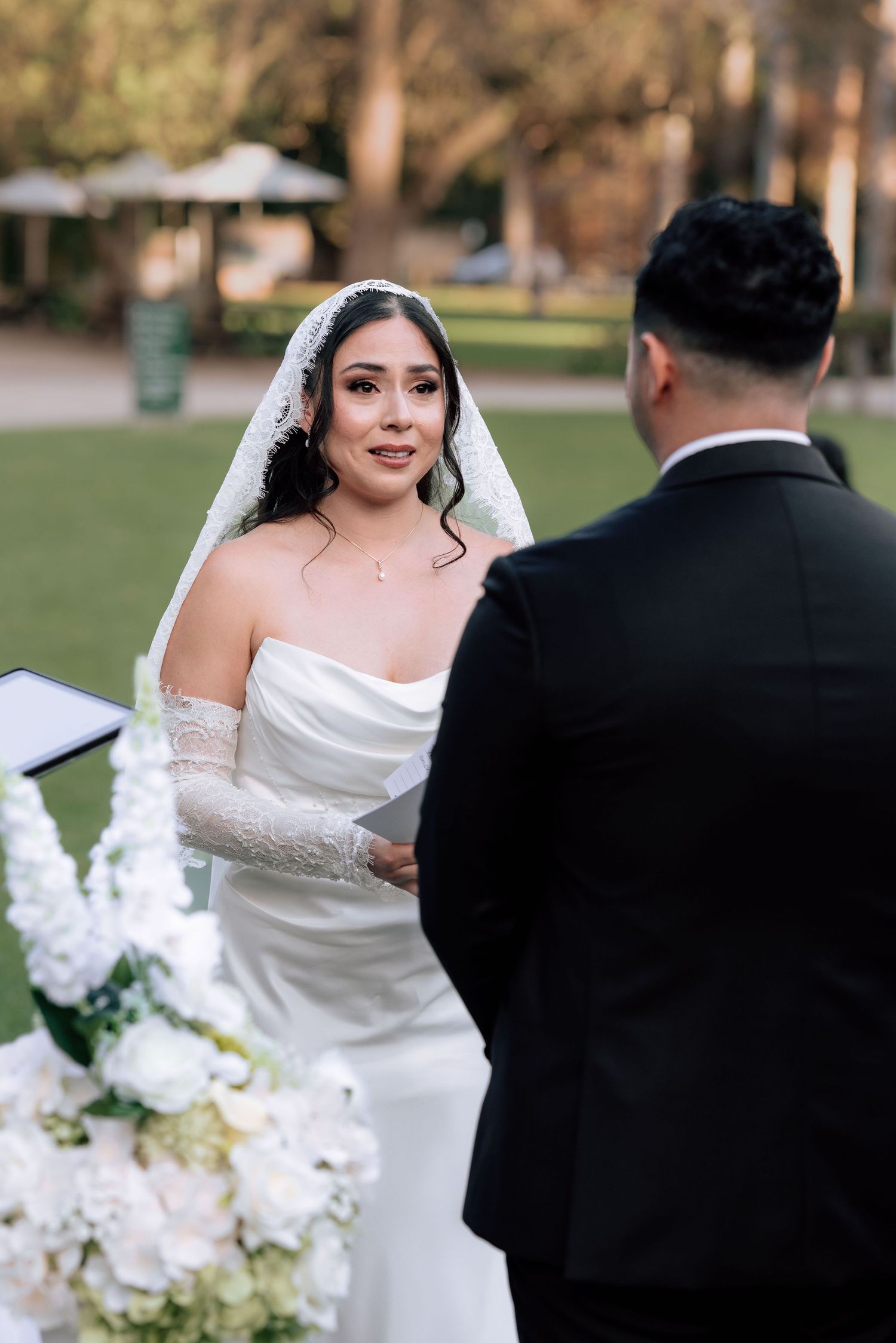 Emotional bride in strapless white gown and lace veil reading vows to groom at outdoor ceremony.