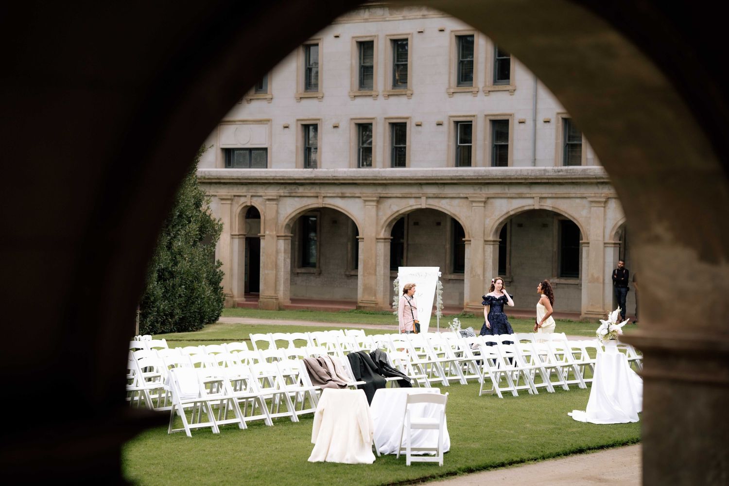 Outdoor wedding ceremony setup with white chairs on lawn viewed through stone archway.