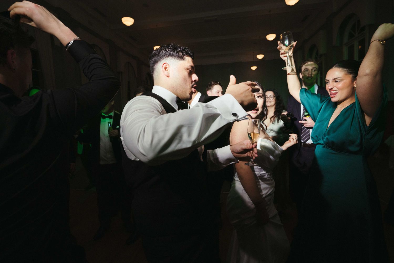 Groom in gray suit dances energetically with guests at lively wedding reception under green ambient lighting.