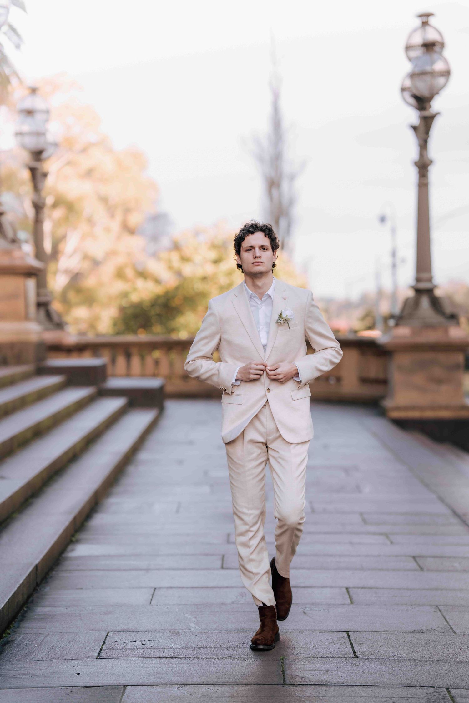 Groom in beige suit adjusting jacket while walking on stone path near vintage lamp posts.