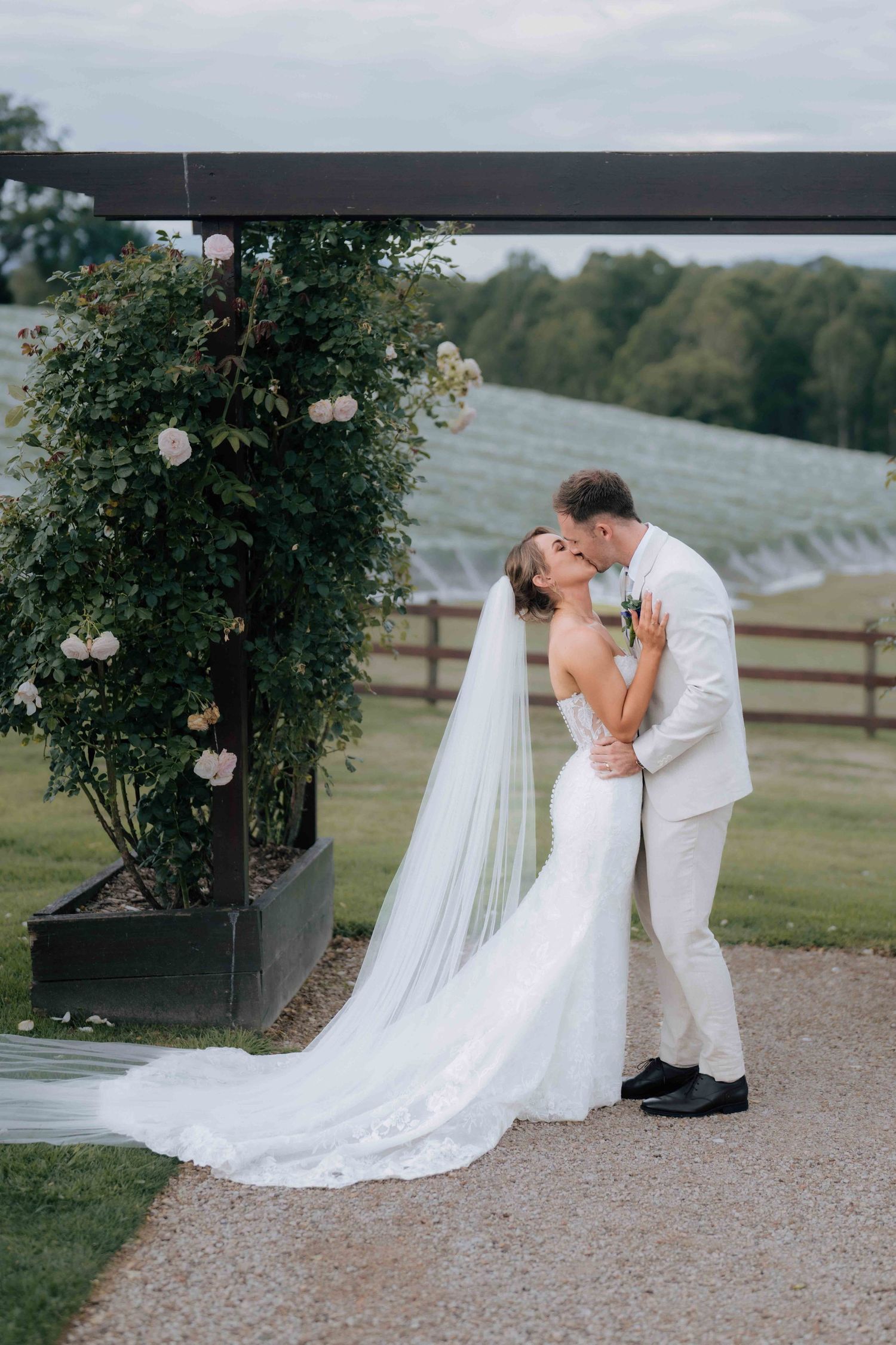 Bride and groom share first kiss at floral arch with vineyard backdrop at outdoor wedding ceremony.