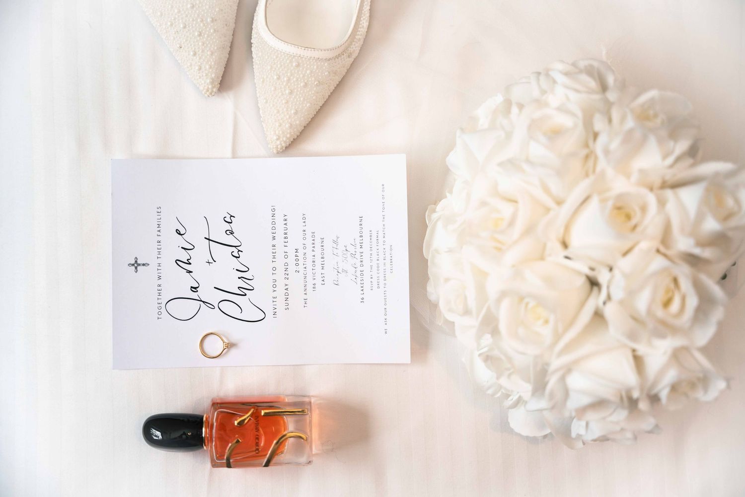 Wedding flat lay featuring invitation, white rose bouquet, perfume bottle, rings, and bridal shoes.