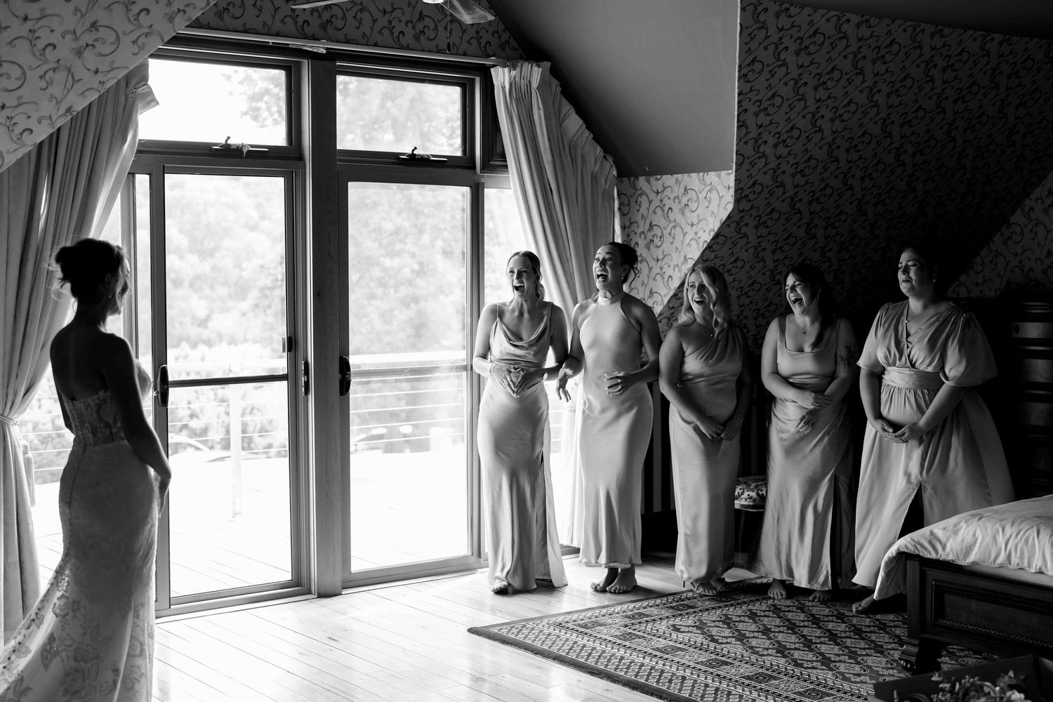 Black and white photo of bridesmaids reacting with joy as bride reveals her wedding dress.