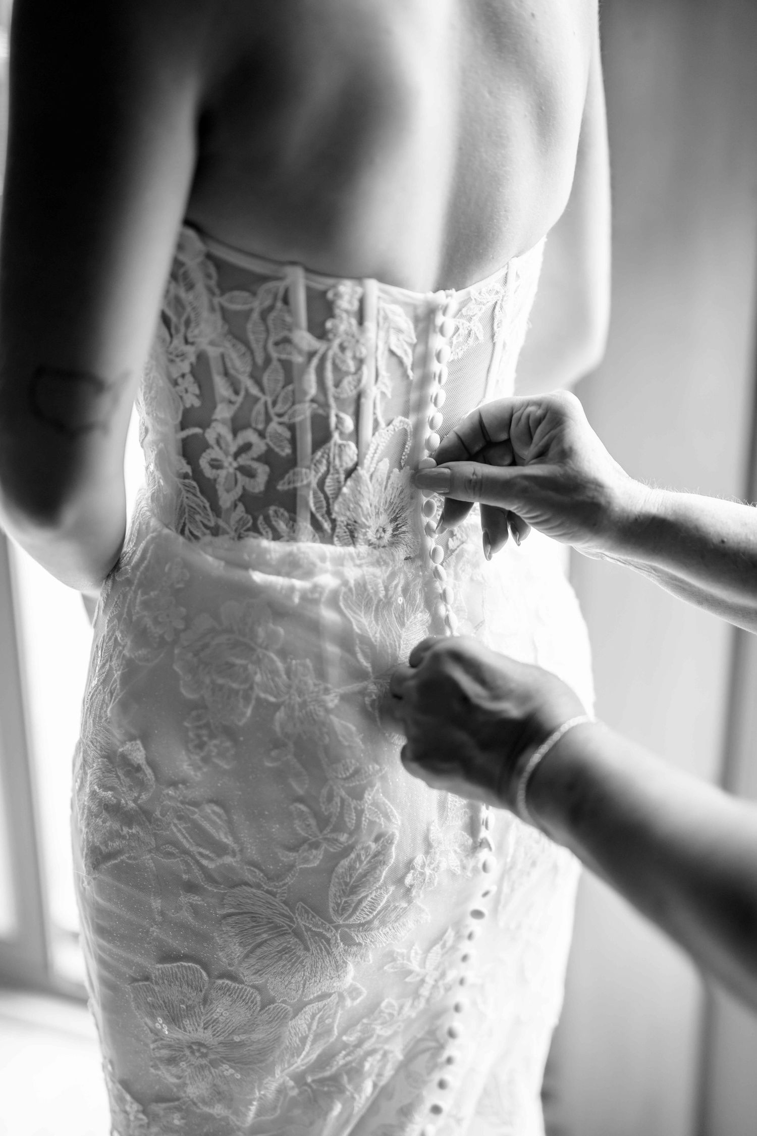 Black and white close-up of bride's lace wedding dress being buttoned from behind.